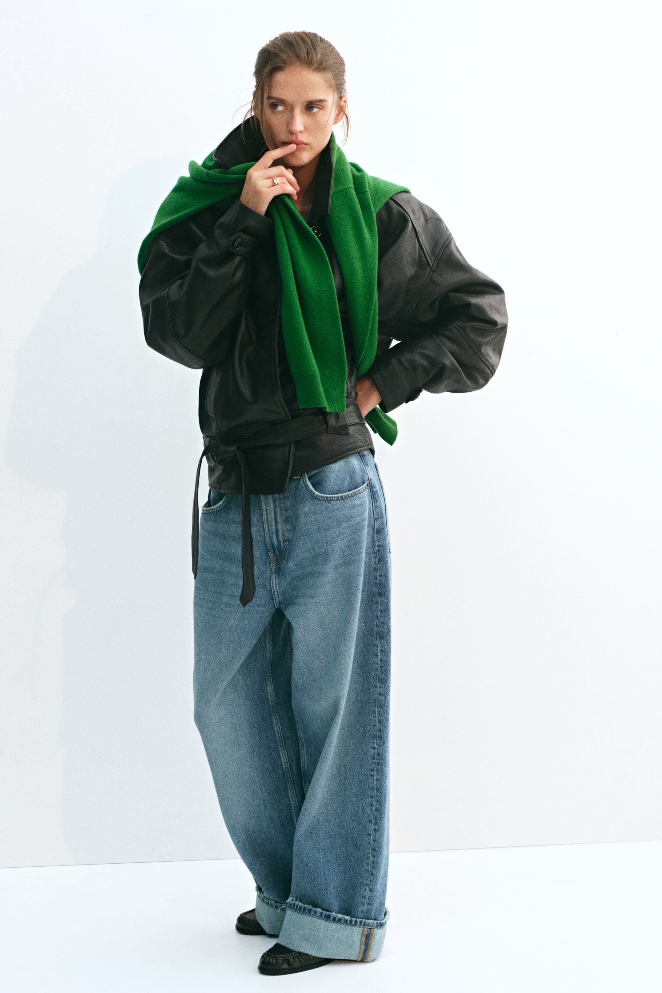 Προβολή μεγαλύτερης εικόνας: Τζιν σε μπλε denim με φαρδύ μπατζάκι, κανονική μέση και χοντρά αναδιπλωμένα ρεβέρ, φορεμένα από το μοντέλο. Το τζιν συνδυάζεται με μαύρο δερμάτινο jacket με ζώνη και πράσινο πλεκτό πουλόβερ ριγμένο στους ώμους.