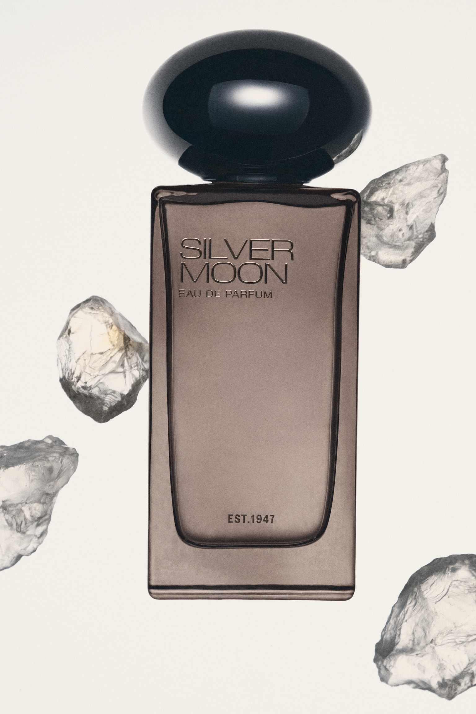 A rectangular perfume bottle with a smoky brown, semi-translucent glass body stands upright, featuring a large, glossy black dome-shaped cap. The front of the bottle is embossed with silver-toned text reading "SILVER MOON EAU DE PARFUM" near the top and "EST.1947" near the bottom. - 1
