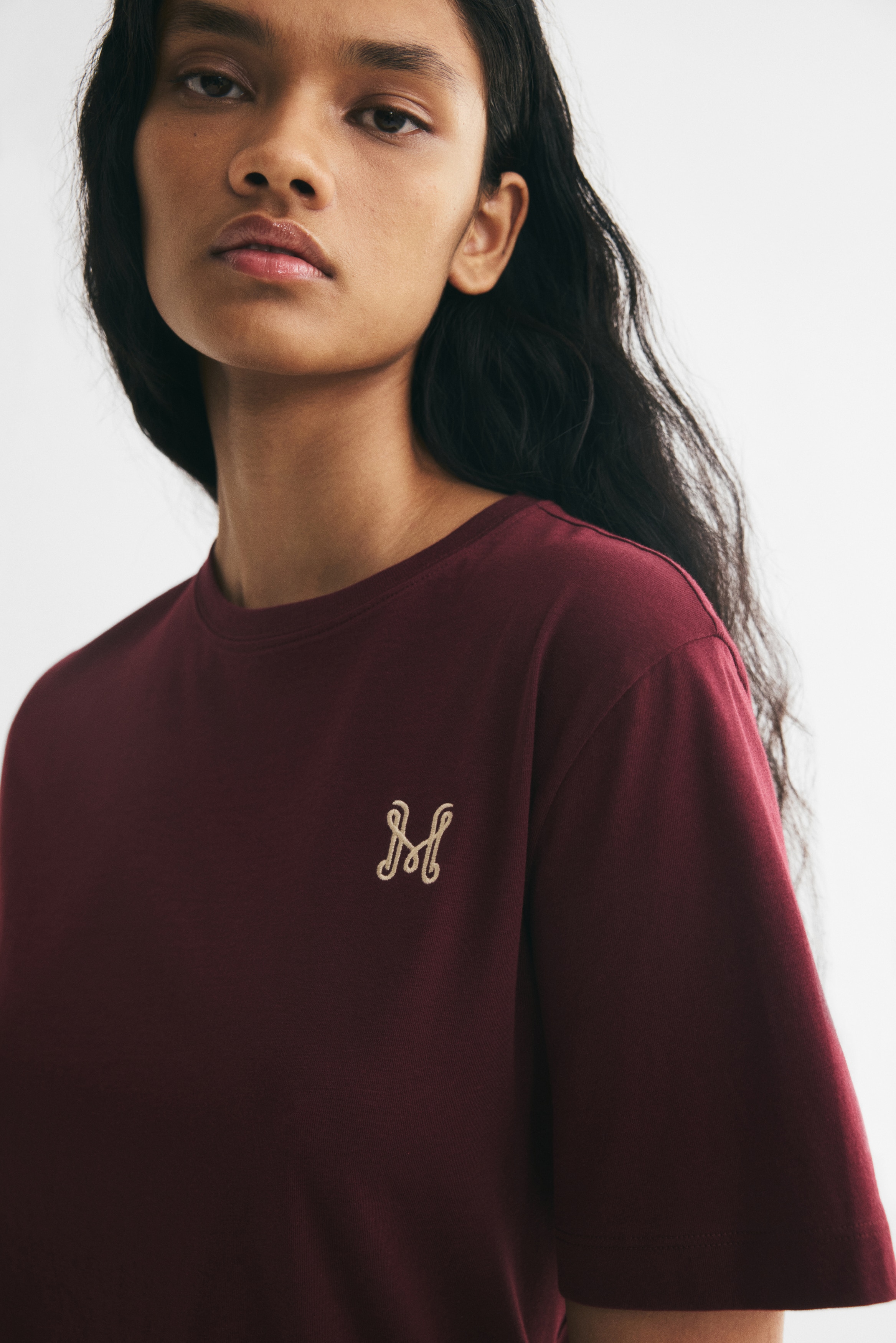 The maroon t-shirt is showcased from the front, highlighting a crew neck and short sleeves. A subtle, cream-coloured 'M' logo is embroidered on the left chest.