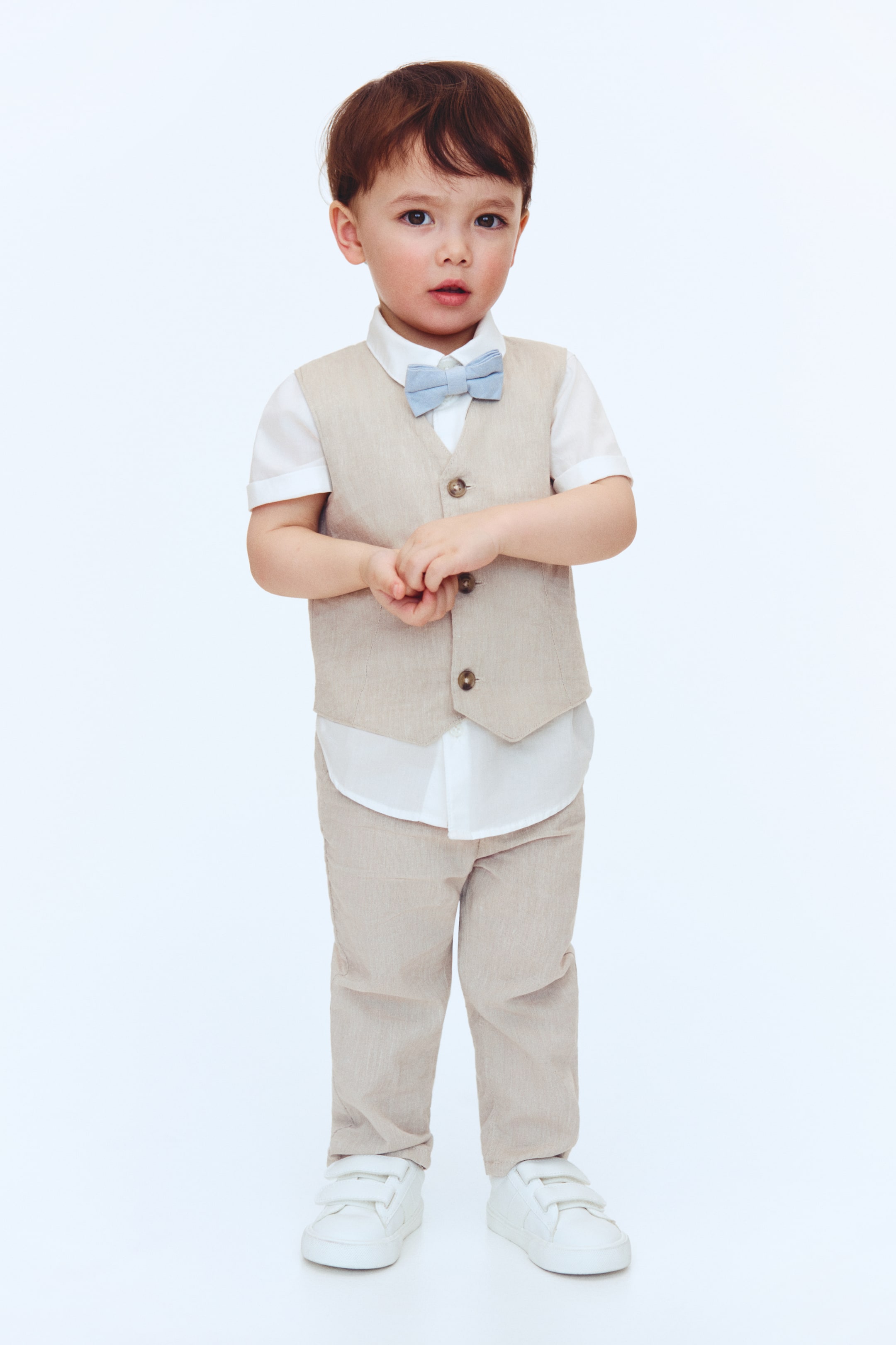 View larger image: A child's beige vest and matching full-length pants set is displayed, layered over a white short-sleeved collared shirt and styled with a light blue bow tie. The vest showcases a V-neckline and three visible brown buttons, while the ensemble is completed with white velcro sneakers, facing forward.