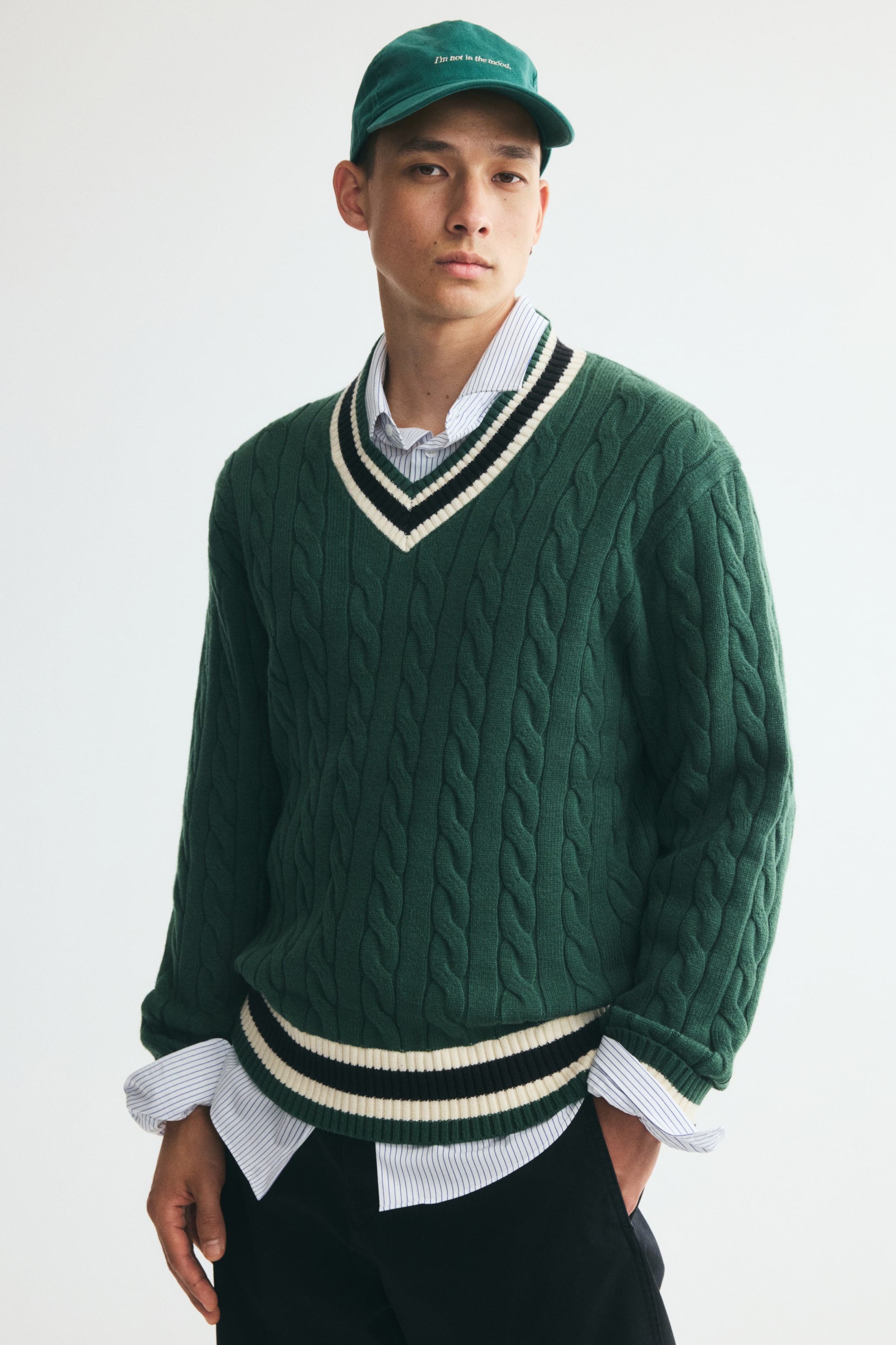 View larger image: A green cable-knit sweater is being worn by the model, layered over a light-colored striped button-down shirt. The sweater features a relaxed fit, a ribbed V-neck with cream and black contrast stripes, and a matching striped ribbed hem.