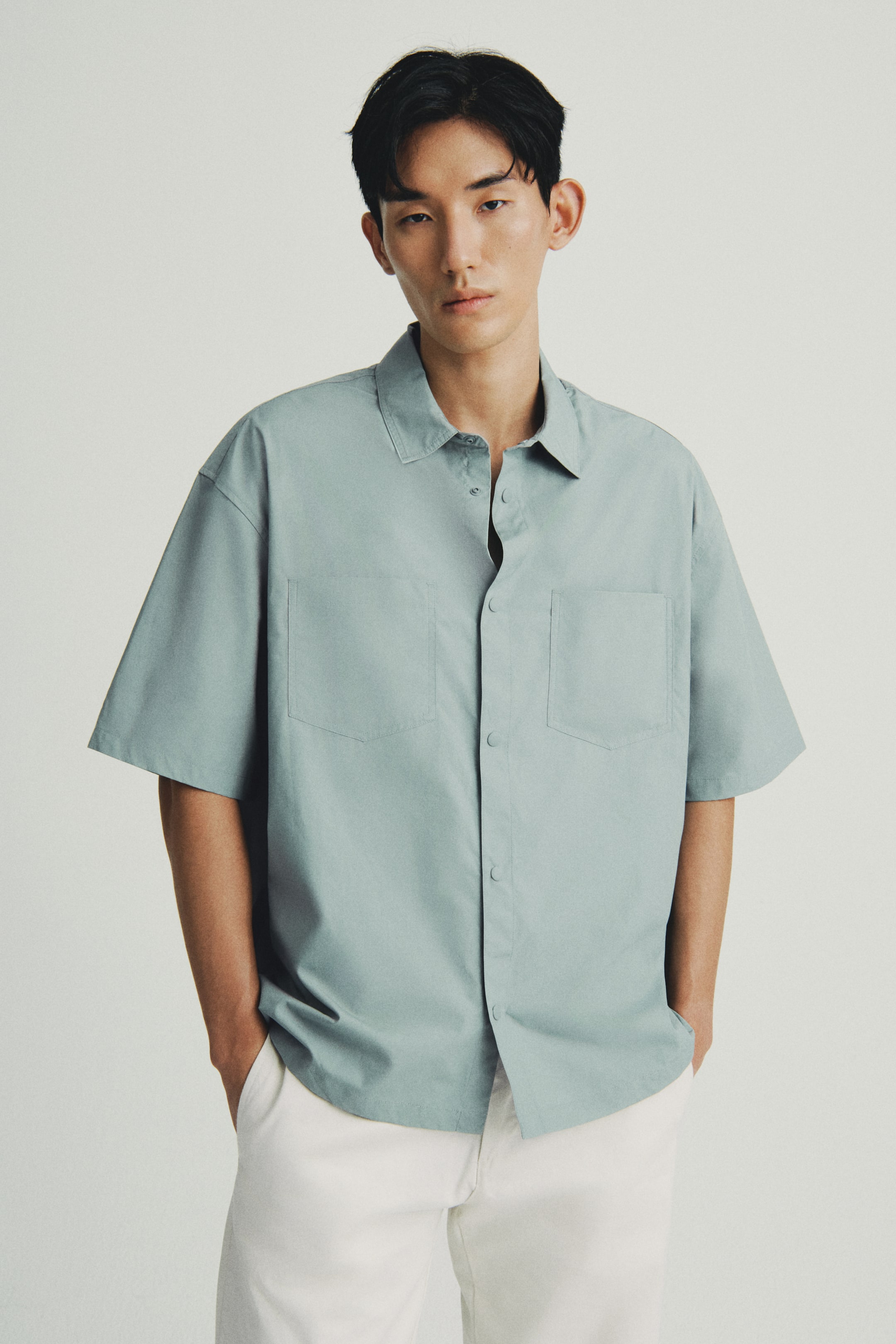 View larger image: The pale blue short-sleeved shirt is being presented front-facing, showcasing a spread collar, a button-front closure with visible buttons, and two patch pockets being displayed on the chest. The shirt is exhibiting a relaxed fit, with its hem falling untucked over white pants.