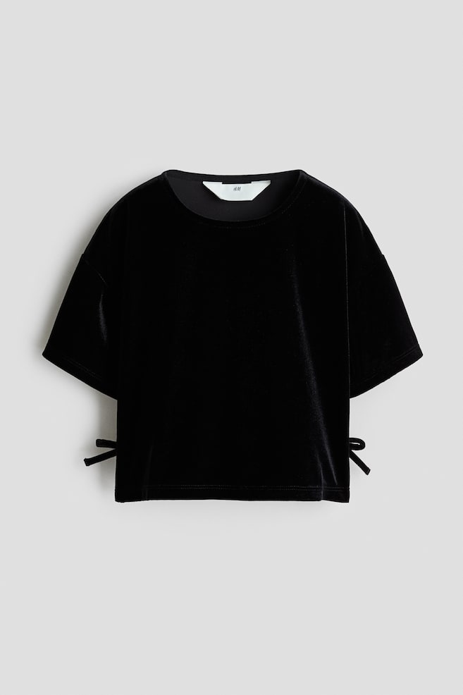 A black velvet crop top is shown from the front, featuring a round neckline, relaxed short sleeves, and small decorative tie details at the lower side seams. - 1