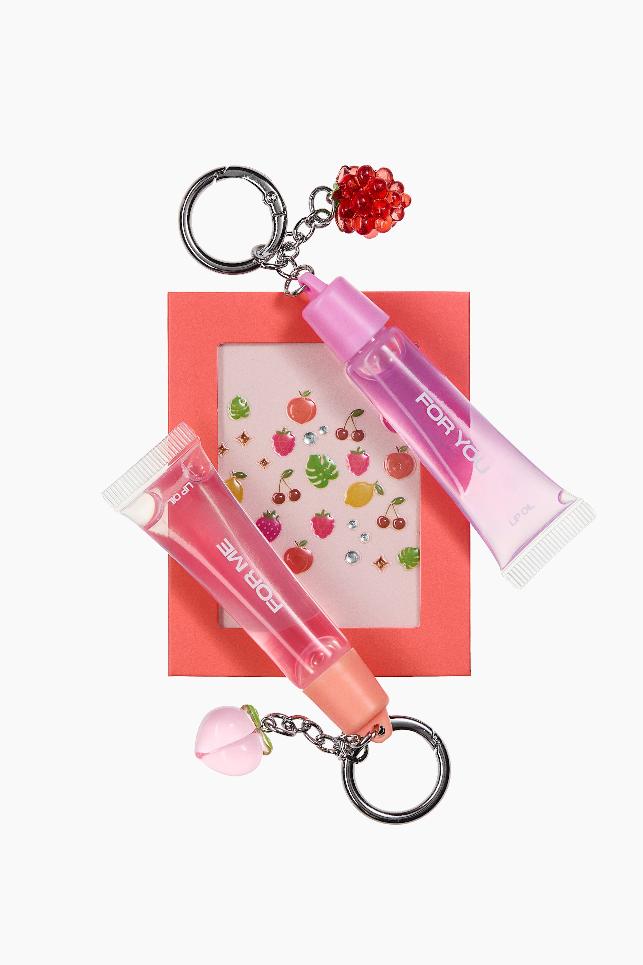 Agrandir l'image: Deux tubes de gloss à lèvres transparents, chacun attaché à un porte-clés argenté, reposent sur une carte carrée de couleur corail ornée de stickers de fruits et de gemmes transparentes. Le tube supérieur présente une teinte rose-violet avec un bouchon rose et un charm framboise rouge perlé, tandis que le tube inférieur arbore une teinte rose-pêche avec un bouchon pêche et un charm pêche rose translucide. Les deux tubes affichent le texte "LIP OIL" et soit "FOR YOU" soit "FOR ME".