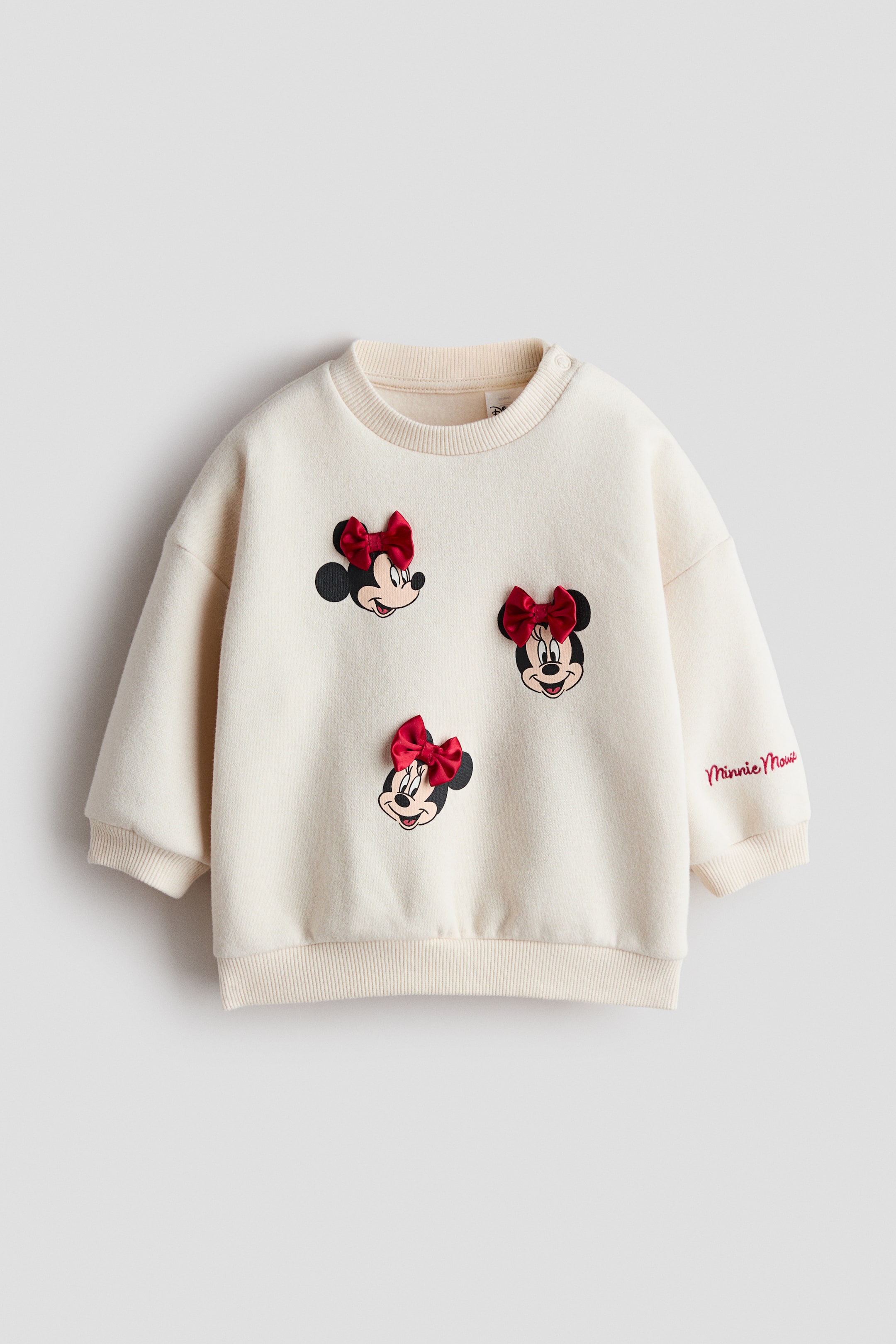 View larger image: The cream-coloured sweatshirt showcases three Minnie Mouse faces adorned with red satin bows appliquéd on the front. It features a ribbed crew neck, snap buttons on the right shoulder, ribbed cuffs, and a ribbed hem. "Minnie Mouse" is embroidered in red script on the left sleeve.
