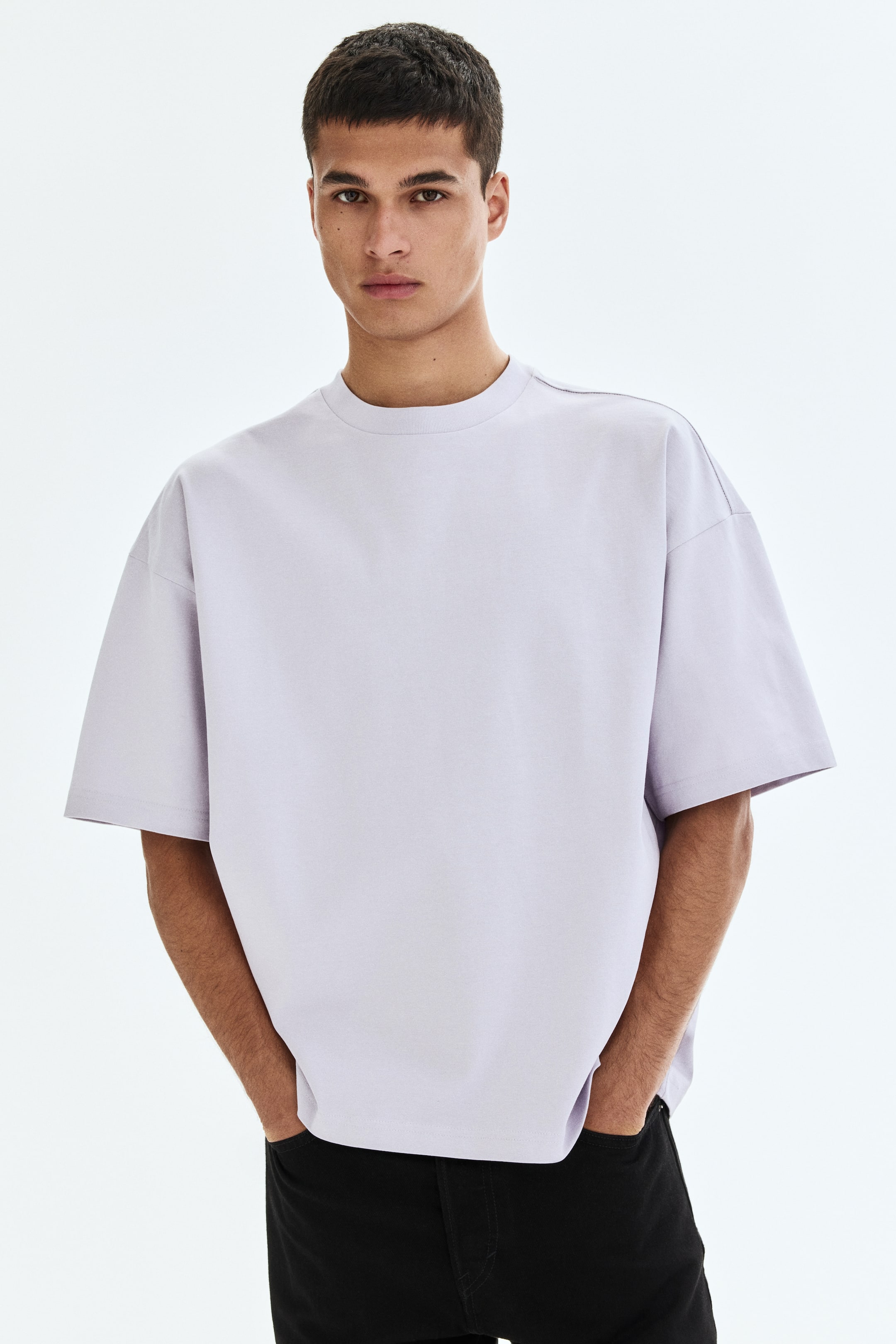 Ver imagen más grande: La modelo lleva puesta una camiseta de estilo cuadrado y holgado en color lila claro, confeccionada en punto de algodón, con un cuello redondo ribeteado, hombros caídos y un dobladillo recto. Las manos de la modelo descansan en los bolsillos de unos pantalones negros, lo que influye en la caída de la camiseta.