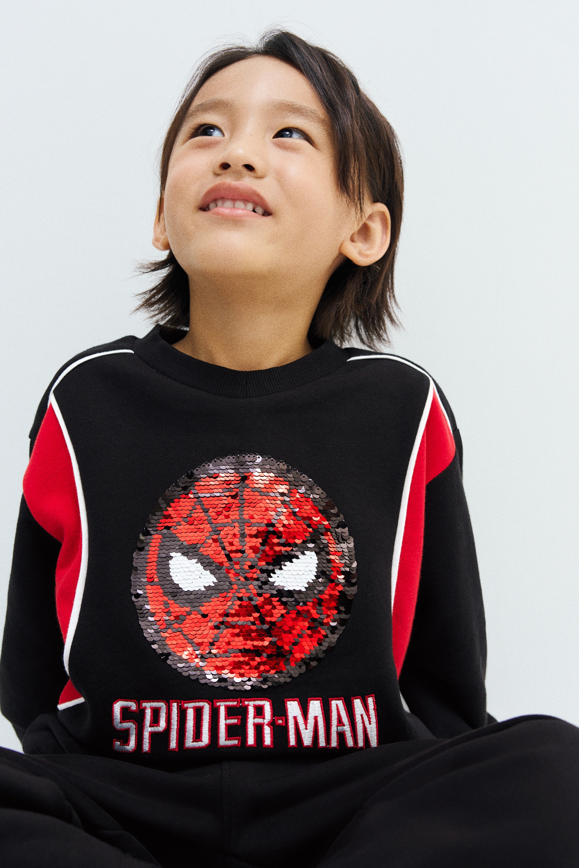 This front-facing black sweatshirt showcases a circular sequined Spider-Man emblem on the chest, with the text "SPIDER-MAN" appearing below in white letters outlined in red. The design features red colour-blocked panels on the shoulders, outlined with white piping, along with a ribbed crew neck, cuffs, and hem.