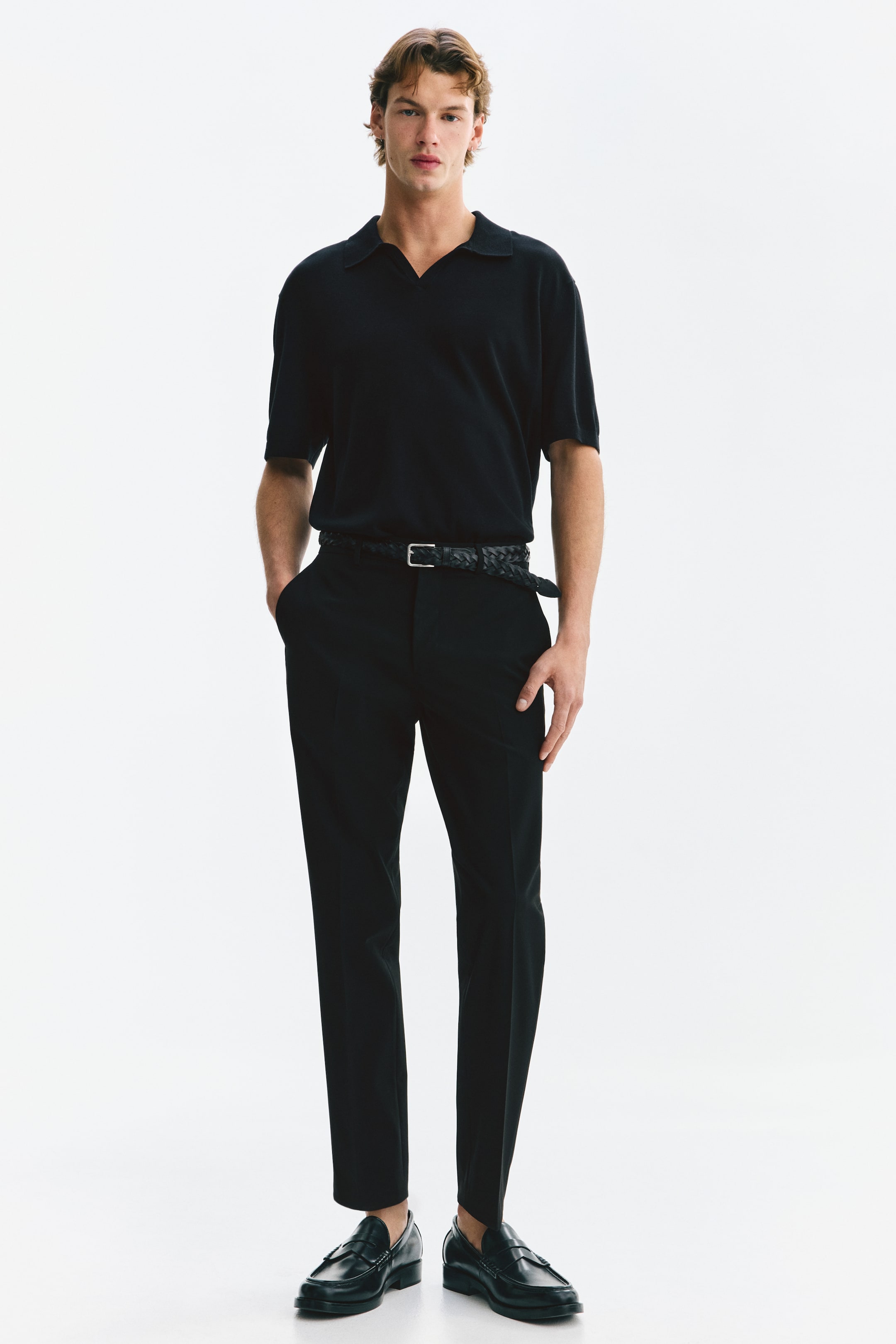 Visualizza l'immagine ingrandita: Un completo interamente nero viene presentato da un'angolazione frontale, caratterizzato da una polo in maglia a maniche corte con colletto aperto, infilata in pantaloni affusolati. Una cintura intrecciata nera con fibbia argentata stringe la vita, mentre mocassini in pelle nera completano il look.