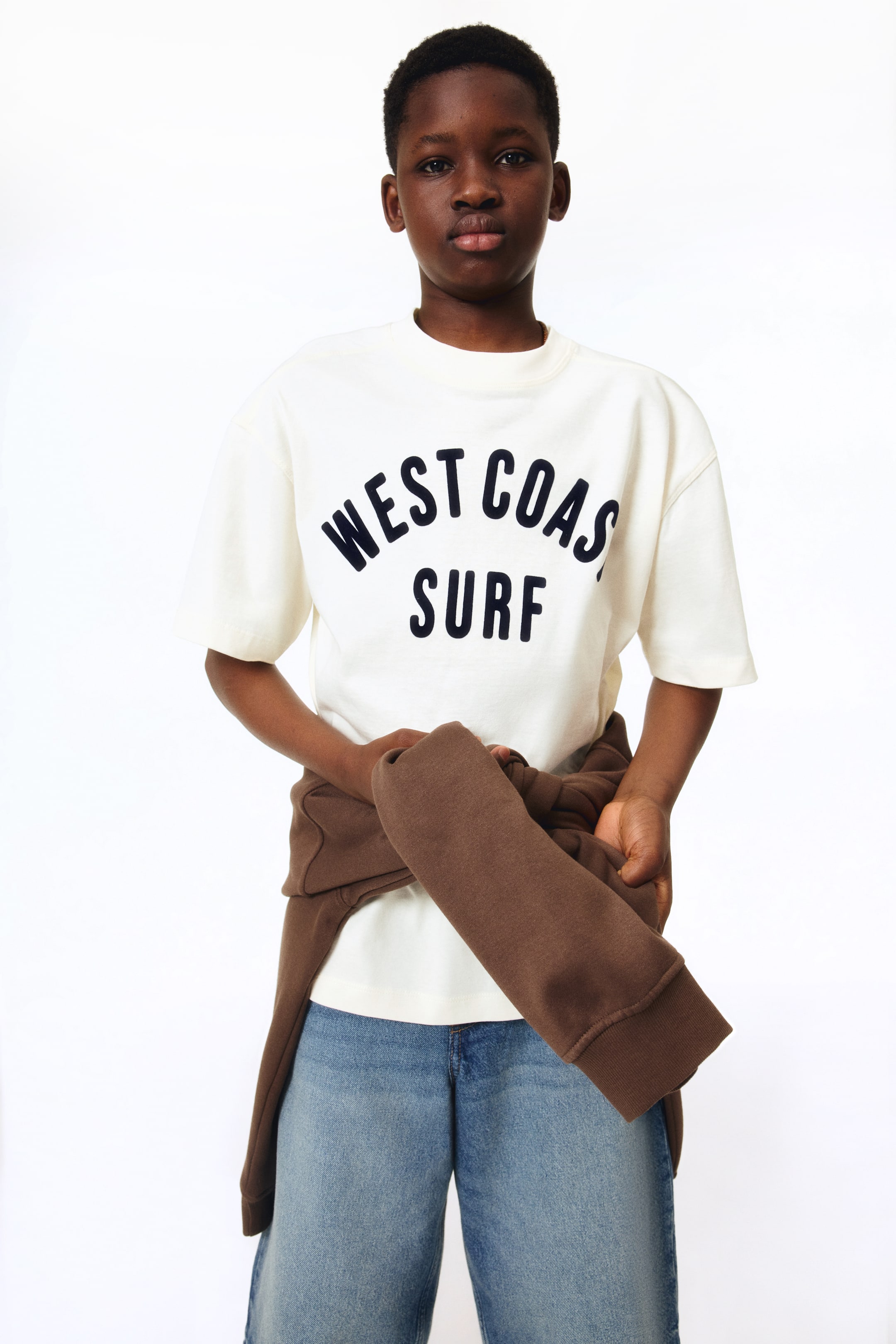 이미지 확대 보기: 루즈핏 크림색 티셔츠는 앞면에 "WEST COAST SURF"라는 어두운 프린트가 특징입니다. 라운드 리브드 넥라인, 드롭 숄더, 짧은 소매, 스트레이트 컷 헴으로 디자인되었습니다.