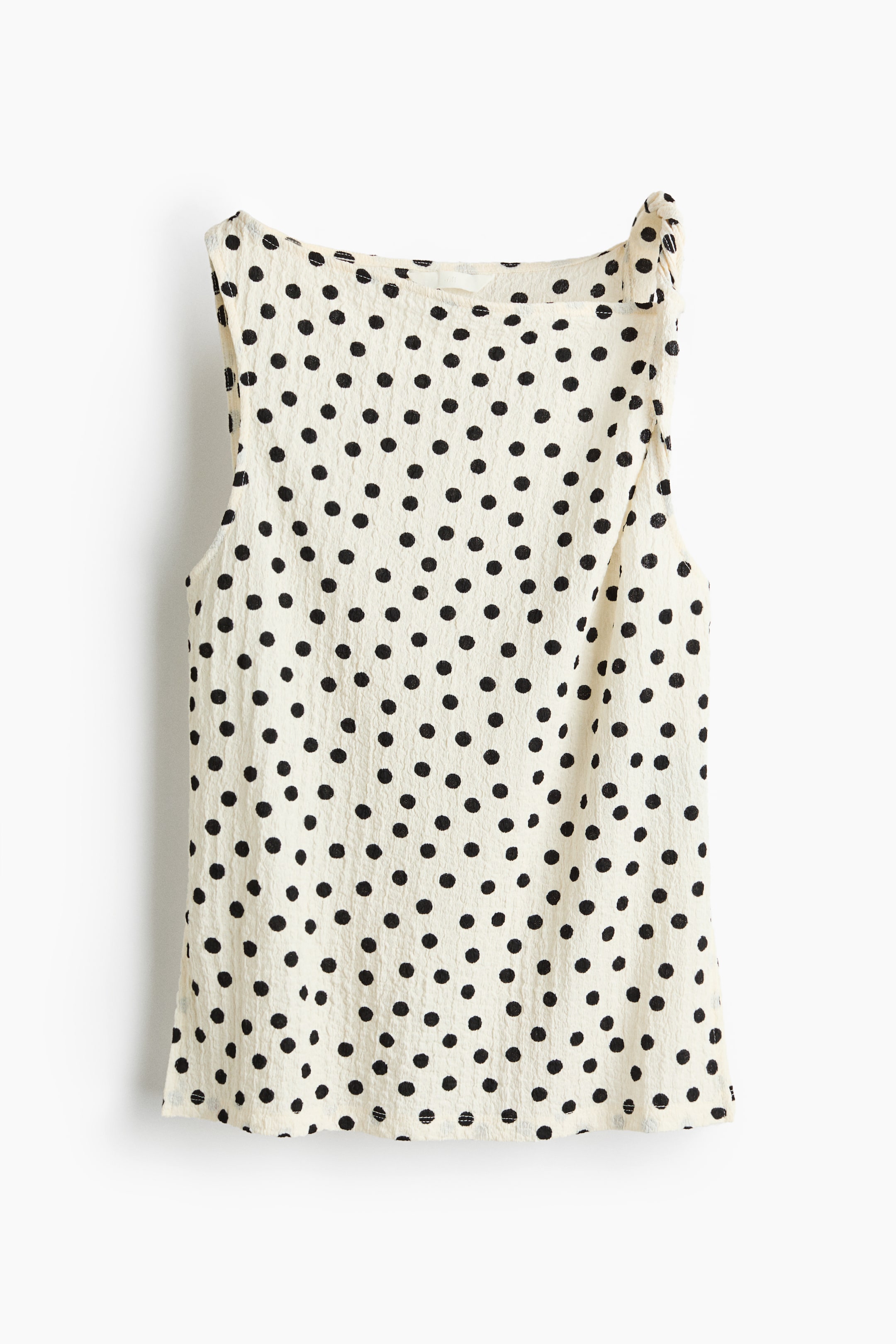 Xem ảnh lớn hơn: Một chiếc áo không tay màu trắng ngà đang hiển thị mẫu chấm bi đen trải dài trên chất liệu vải nhăn, được trình bày từ phía trước. Nó có đặc điểm là cổ áo rộng và chi tiết xếp nếp trên dây vai bên phải.