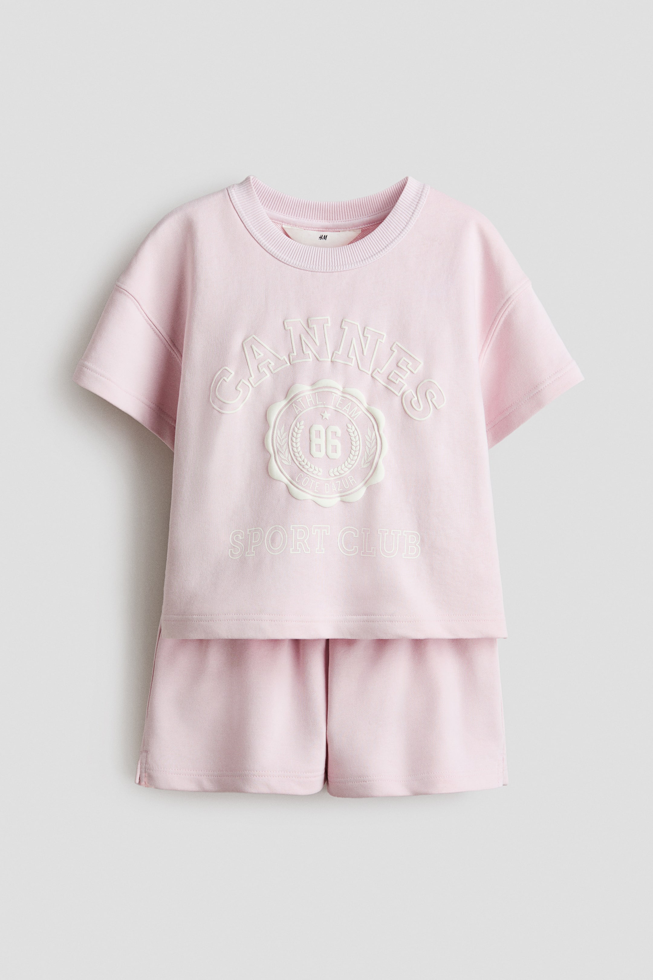 Grösseres Bild ansehen: Ein rosa T-Shirt und Shorts-Set aus bedrucktem, leichtem Sweatshirtstoff wird von vorne präsentiert. Das T-Shirt verfügt über einen gerippten Ausschnitt, überschnittene Schultern und einen erhabenen 'CANNES 86 SPORT CLUB'-Print, während die Shorts einen elastischen Bund mit Kordelzug und einen kleinen Schlitz an einer Seite des Saums aufweisen.