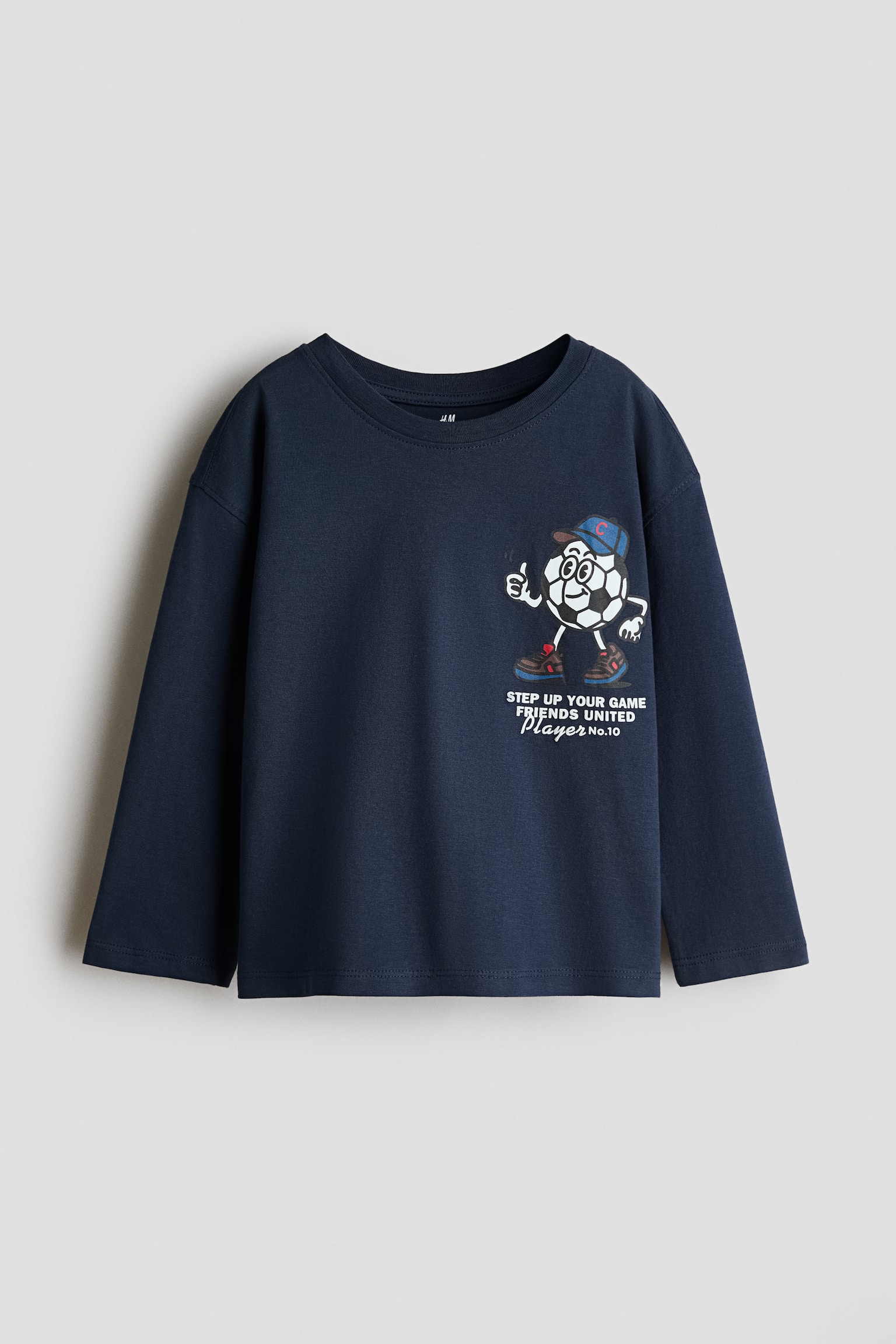 A navy blue long-sleeved cotton jersey T-shirt is shown from the front, featuring dropped shoulders and a ribbed crew neckline. A graphic print on the left chest depicts a cartoon football character with text reading 'STEP UP YOUR GAME FRIENDS UNITED Player No.10.'