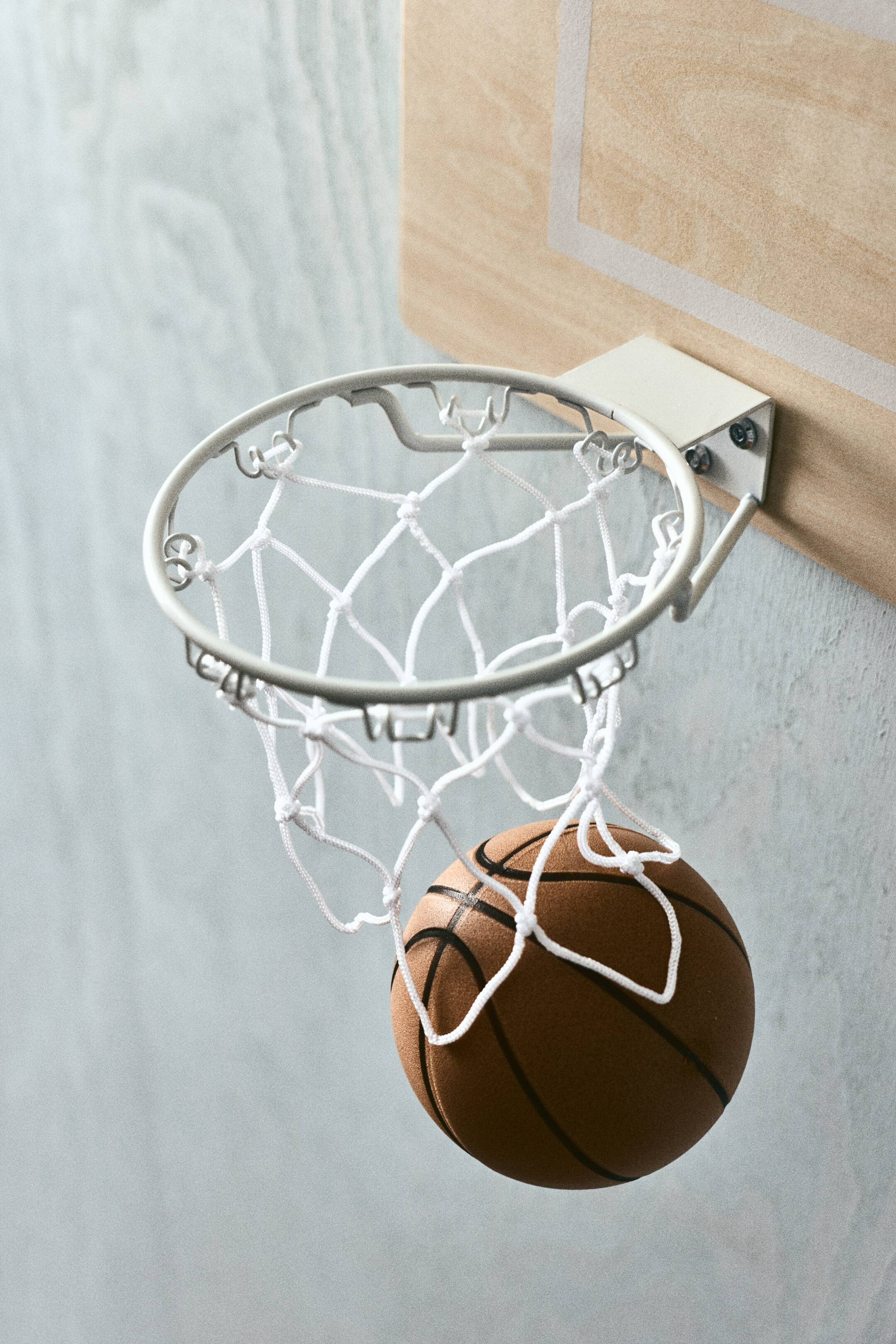 View larger image: A mini basketball hoop featuring a white metal rim and a white net, with a small brown basketball passing through the net. The hoop is securely attached to a light wooden backboard that displays subtle rectangular markings.