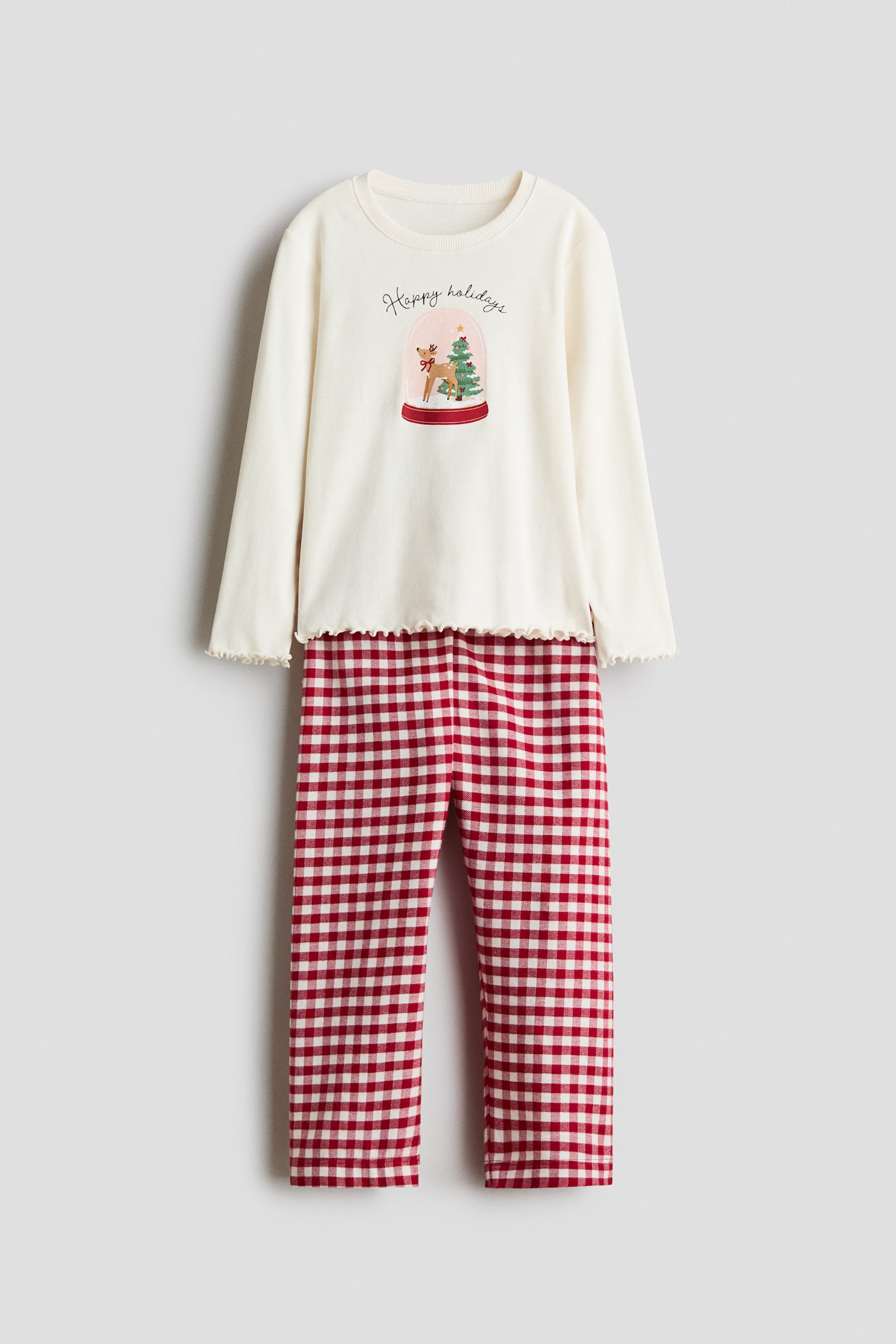 Un set de pijama este prezentat din faÈÄ, format dintr-un top crem cu mÃ¢neci lungi, cu manÈete Èi tiv cu finisaj tip âlettuce edgeâ, Èi un grafic central cu glob de zÄpadÄ Èi textul âHappy holidaysâ. Topul este asortat cu pantaloni drepÈi, cu imprimeu gingham Ã®n carouri roÈii Èi albe.