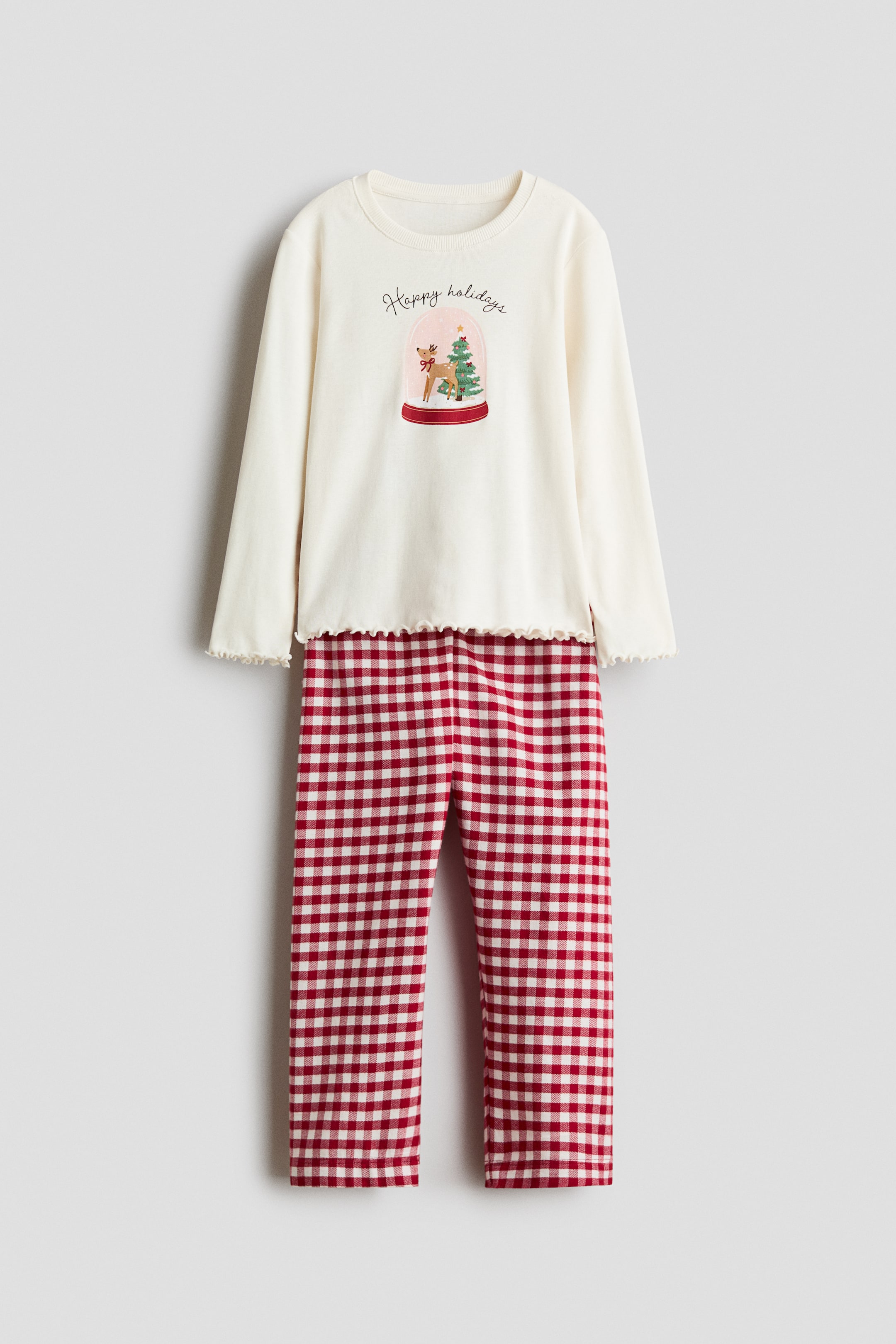 Agrandir l'image: Un ensemble de pyjama est présenté de face, avec un haut crème à manches longues doté de poignets et d’un ourlet à bord ondulé, ainsi qu’un motif central de boule à neige « Happy holidays ». Le haut est assorti à un pantalon droit affichant un motif vichy à carreaux rouges et blancs.