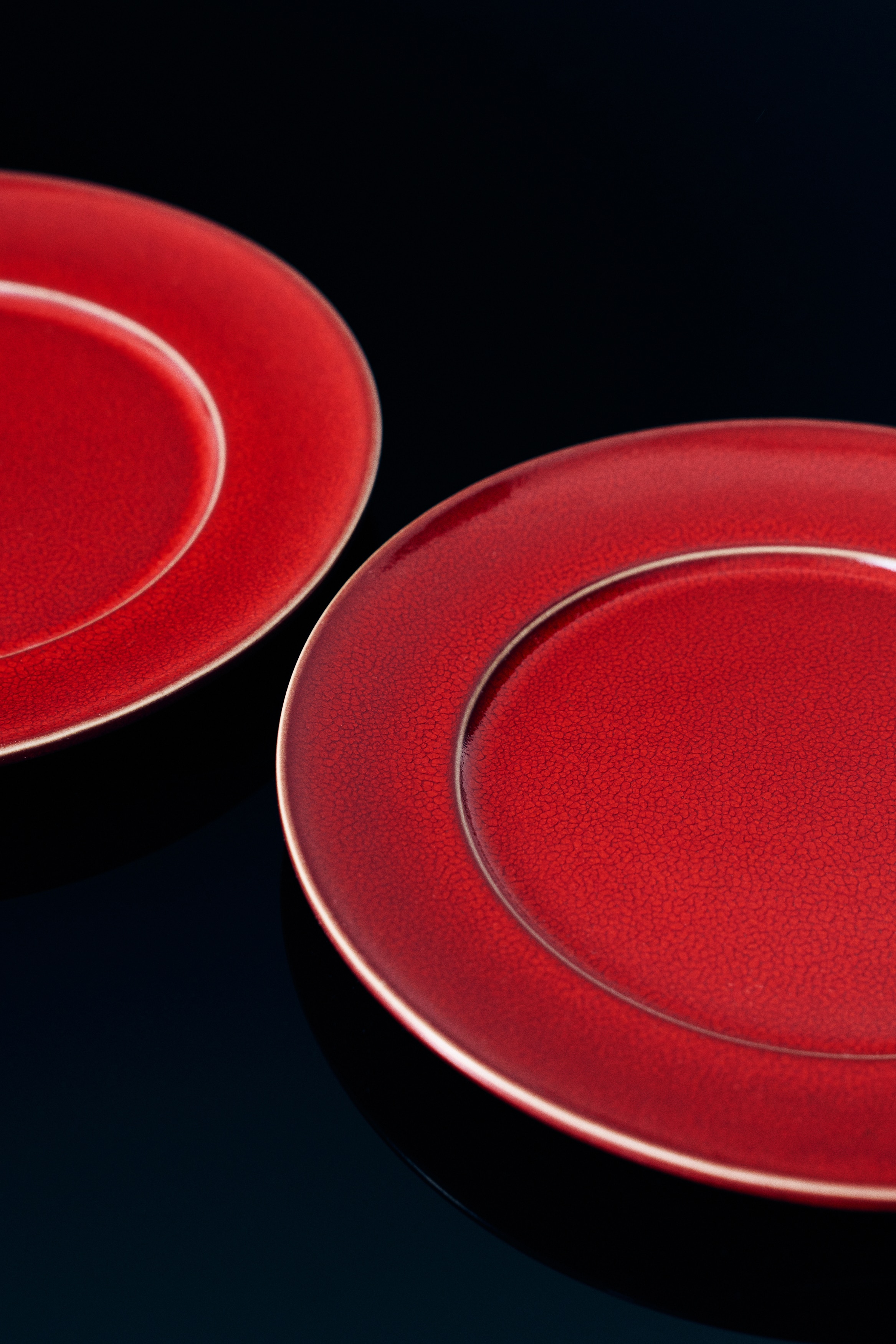 Deux assiettes rondes en céramique présentent un émaillage brillant d’un rouge profond, avec une finition subtilement texturée, vues de dessus. Chaque assiette est dotée d’un rebord surélevé souligné par une fine bordure brun foncé.