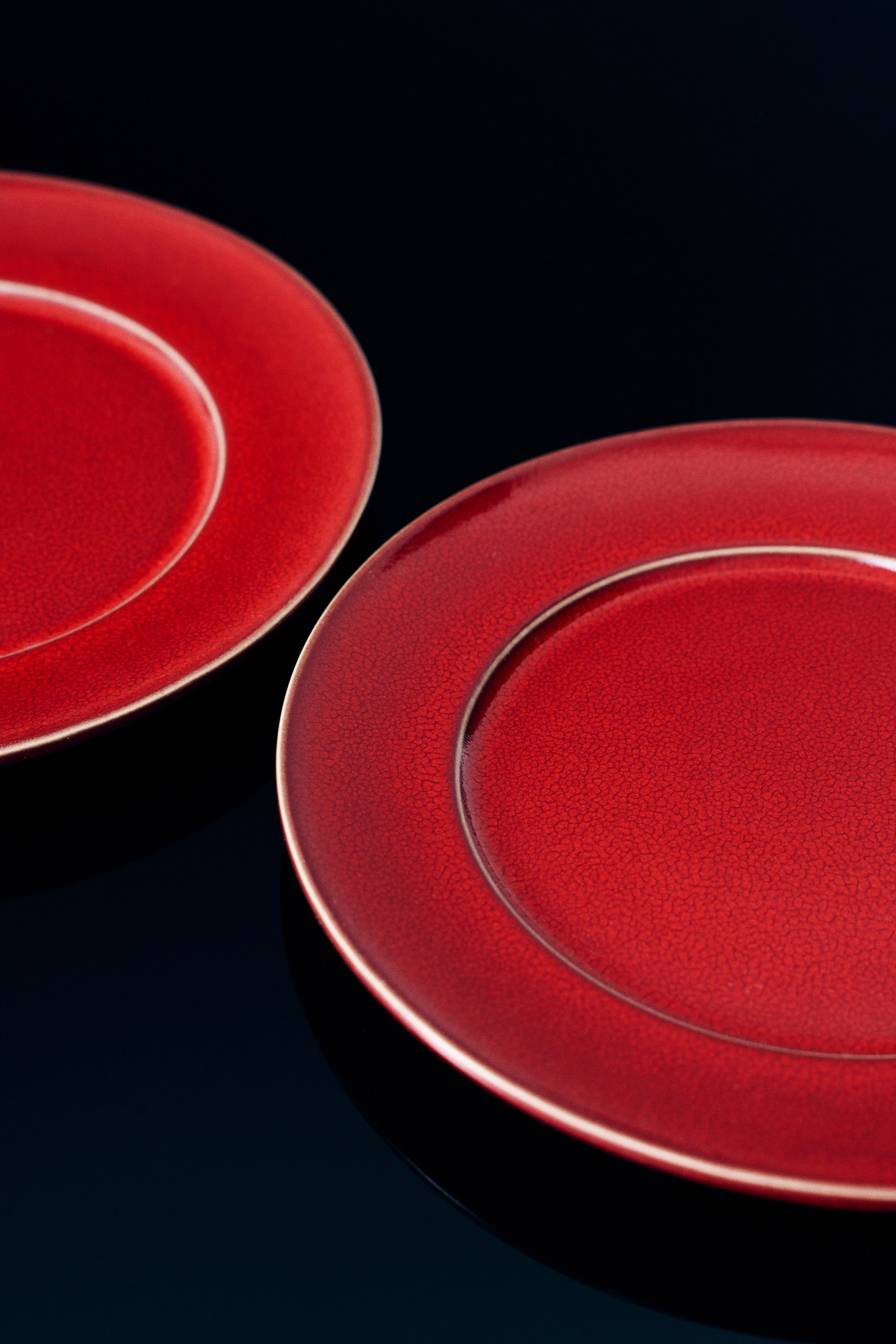 View larger image: Red ceramic plates are shown from an angled top-down view, featuring a vibrant red glossy finish with a distinctive crackle glaze texture. The plates have a raised rim and a recessed centre well, with a thin, lighter-toned edge highlighting their circular shape.
