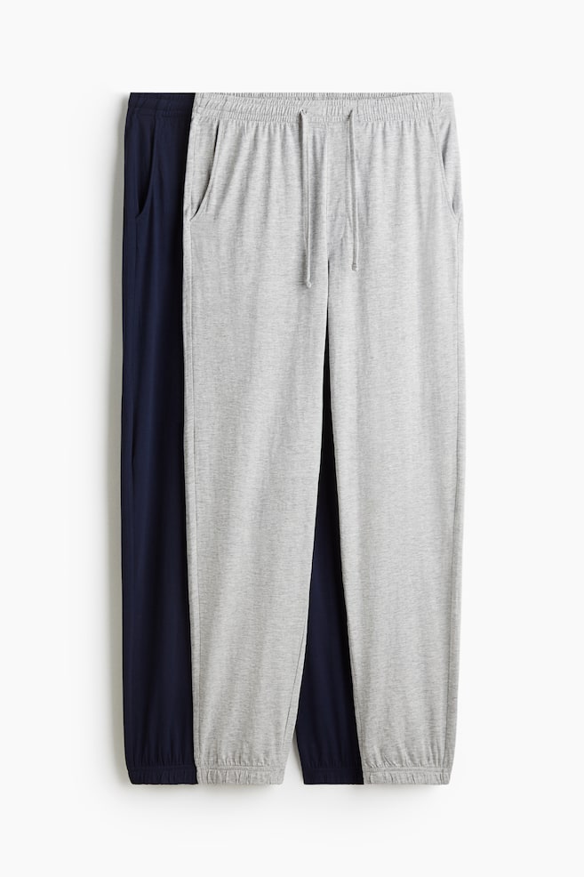 Two pairs of cotton jersey sweatpants are presented facing FRONT. The light gray pair, positioned in front, is featuring an elasticated, drawstring waist, a stitched fly detail, side pockets, and ribbed hems. A dark blue pair is partially visible behind it, also displaying side pockets and ribbed hems. - 1