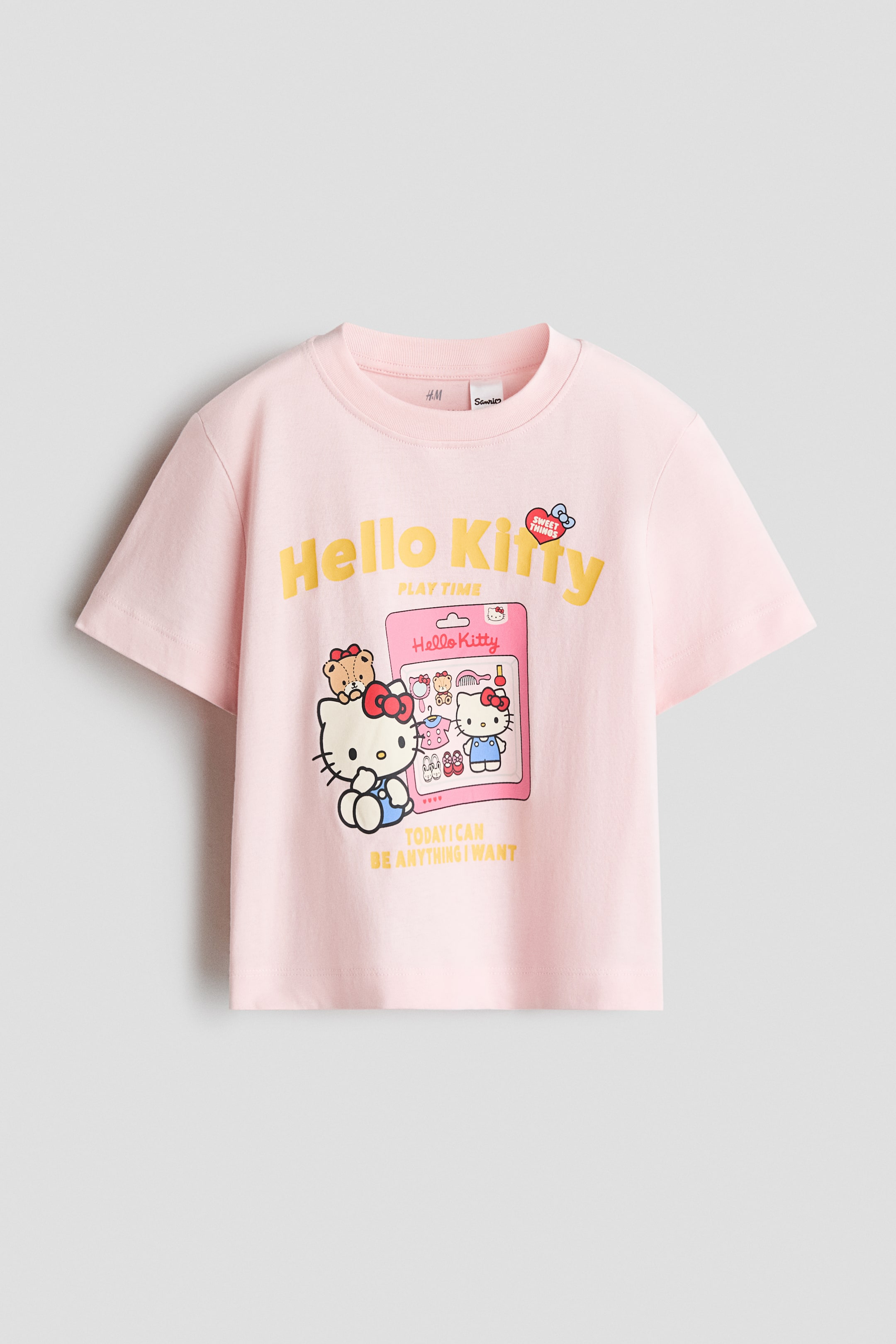View larger image: A pink short-sleeve t-shirt features a crew neck and a graphic print on the front. The print showcases "Hello Kitty" in yellow text, a seated Hello Kitty figure with a teddy bear, and a pink toy package illustration depicting another Hello Kitty with accessories and the phrase "Today I can be anything I want."