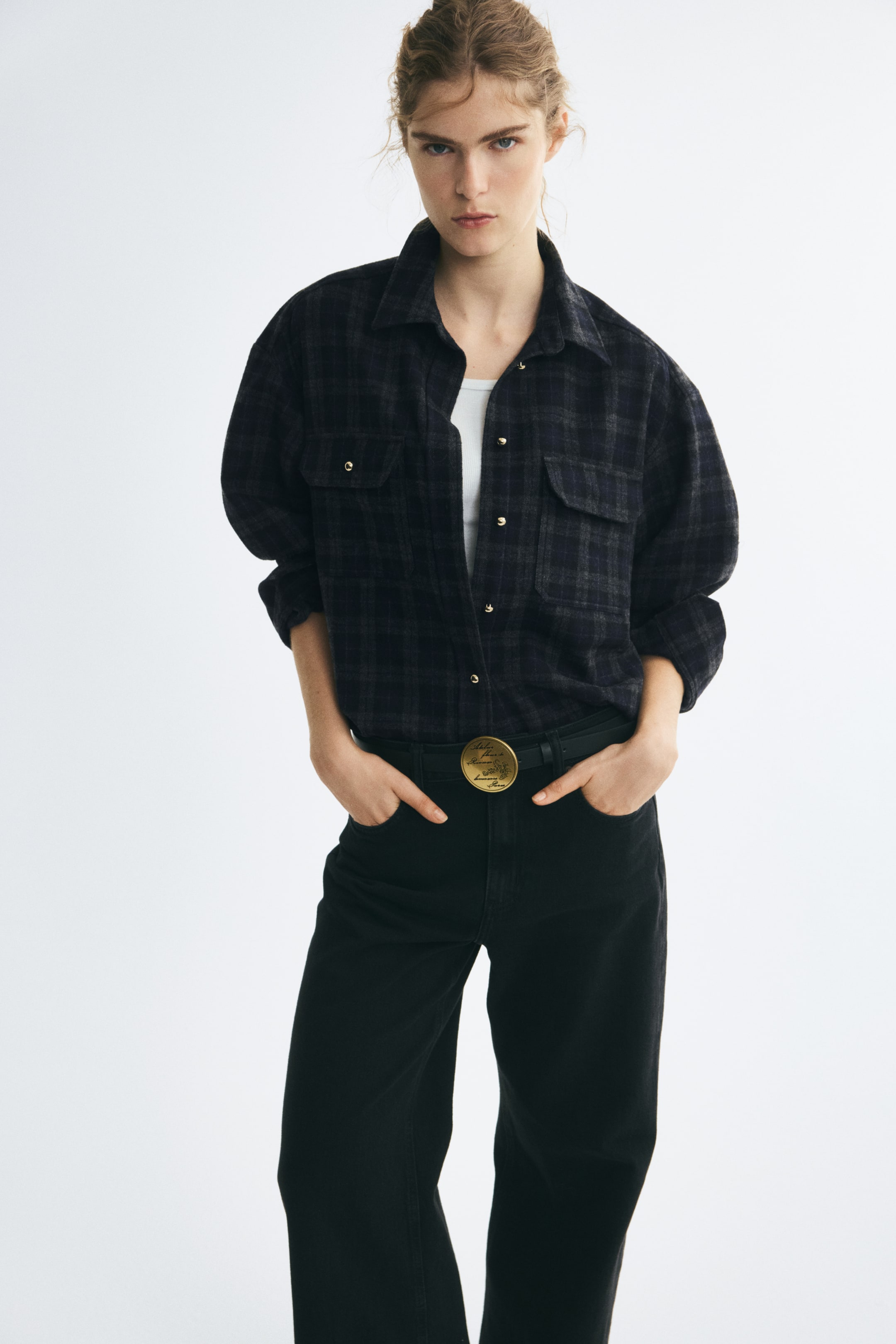 Ampliar la imagen: La modelo lleva una camisa de franela a cuadros de corte oversize en un patrón de cuadros oscuros, combinada sobre un top blanco y metida dentro de pantalones negros. La camisa presenta cuello, botones en tono dorado al frente, hombros caídos y bolsillos de parche en el pecho con tapas abotonadas.
