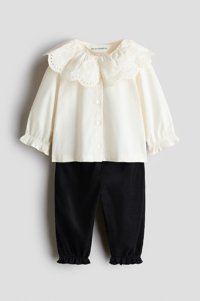 Ein zweiteiliges Kinderoutfit zeigt eine Frontansicht einer cremefarbenen Bluse, kombiniert mit einer schwarzen Cordhose. Die Bluse verfügt über einen groÃen, gewellten Kragen mit zarter Stickerei, vorn angeordneten Knopfverschlüssen und gerafften Manschetten, während die Hose elastische, geraffte Beinabschlüsse aufweist. - 1