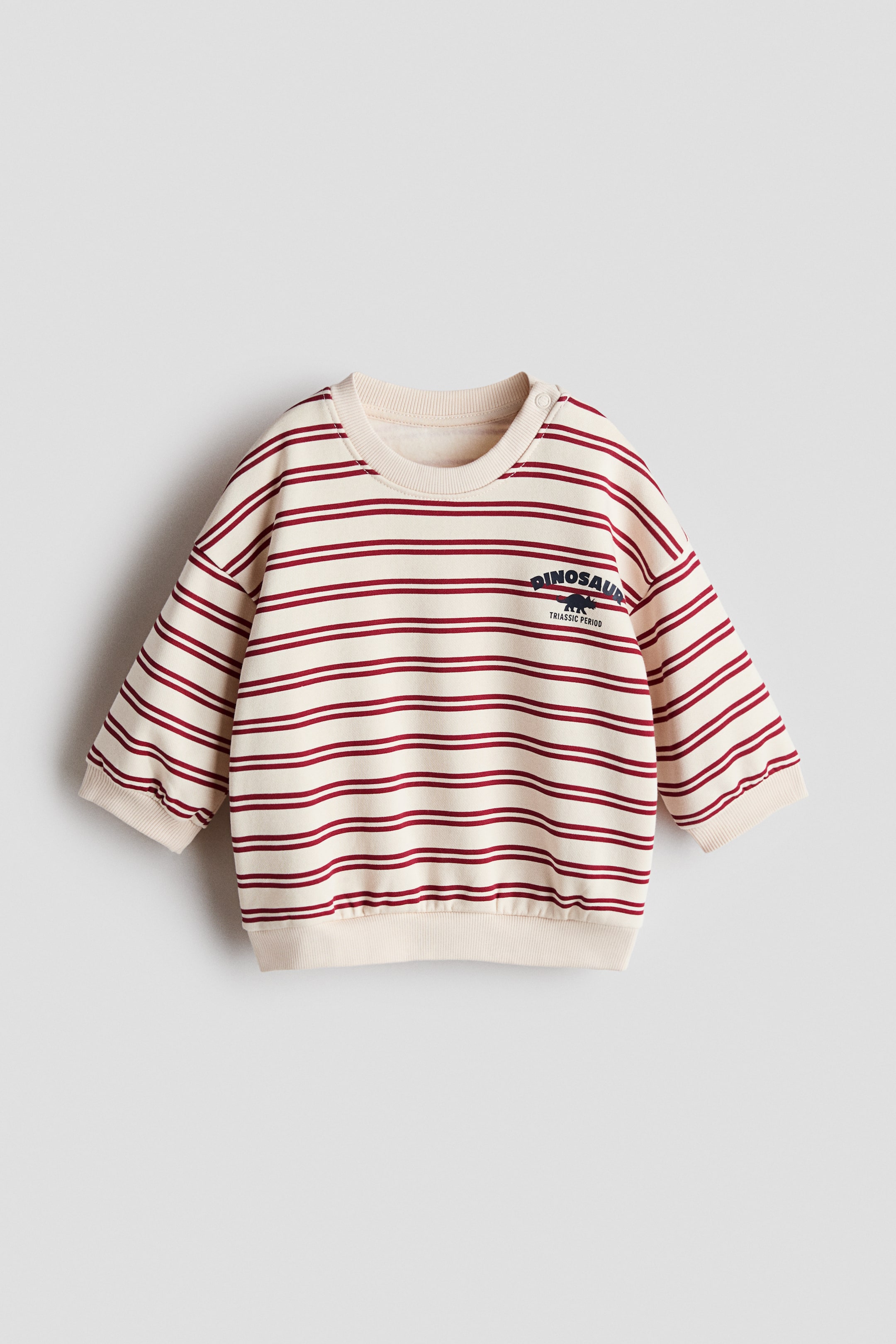 拡大画像を表示: プリント入りのスウェットは正面を向いており、クリーム色のベースに水平な赤いストライプが特徴です。クルーネックで左肩に2つの白いスナップボタンがあり、上胸部には恐竜のシルエットが描かれた濃紺の「DINOSAUR TRIASSIC PERIOD」というグラフィックプリントがあり、リブ付きの袖口と裾が備わっています。