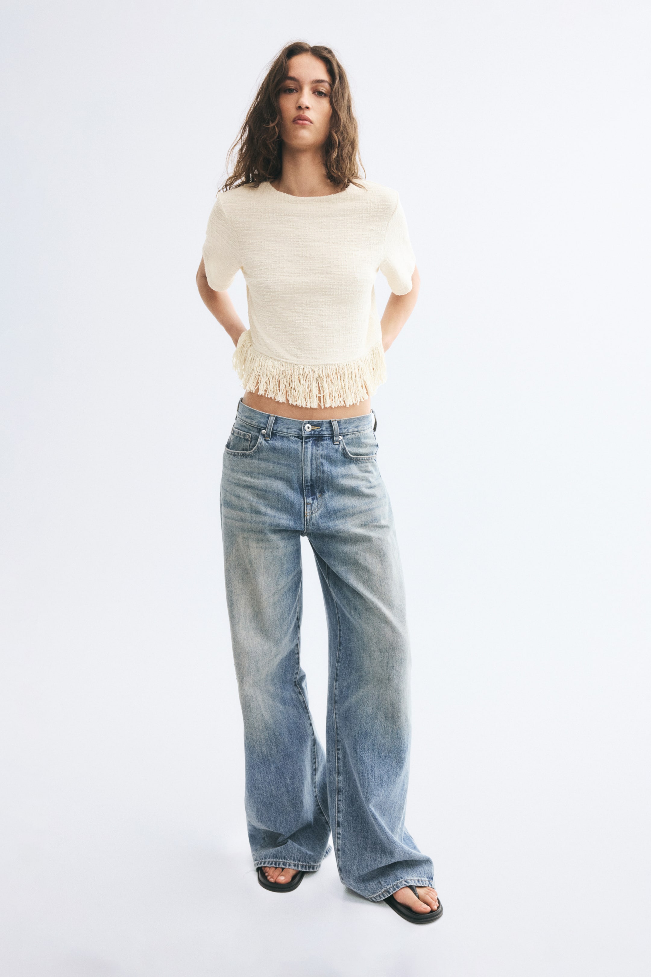 View larger image: A cream-coloured, textured short-sleeve crop top with a curved, fringed hem is worn by the model, paired with wide-leg, light-wash blue jeans.