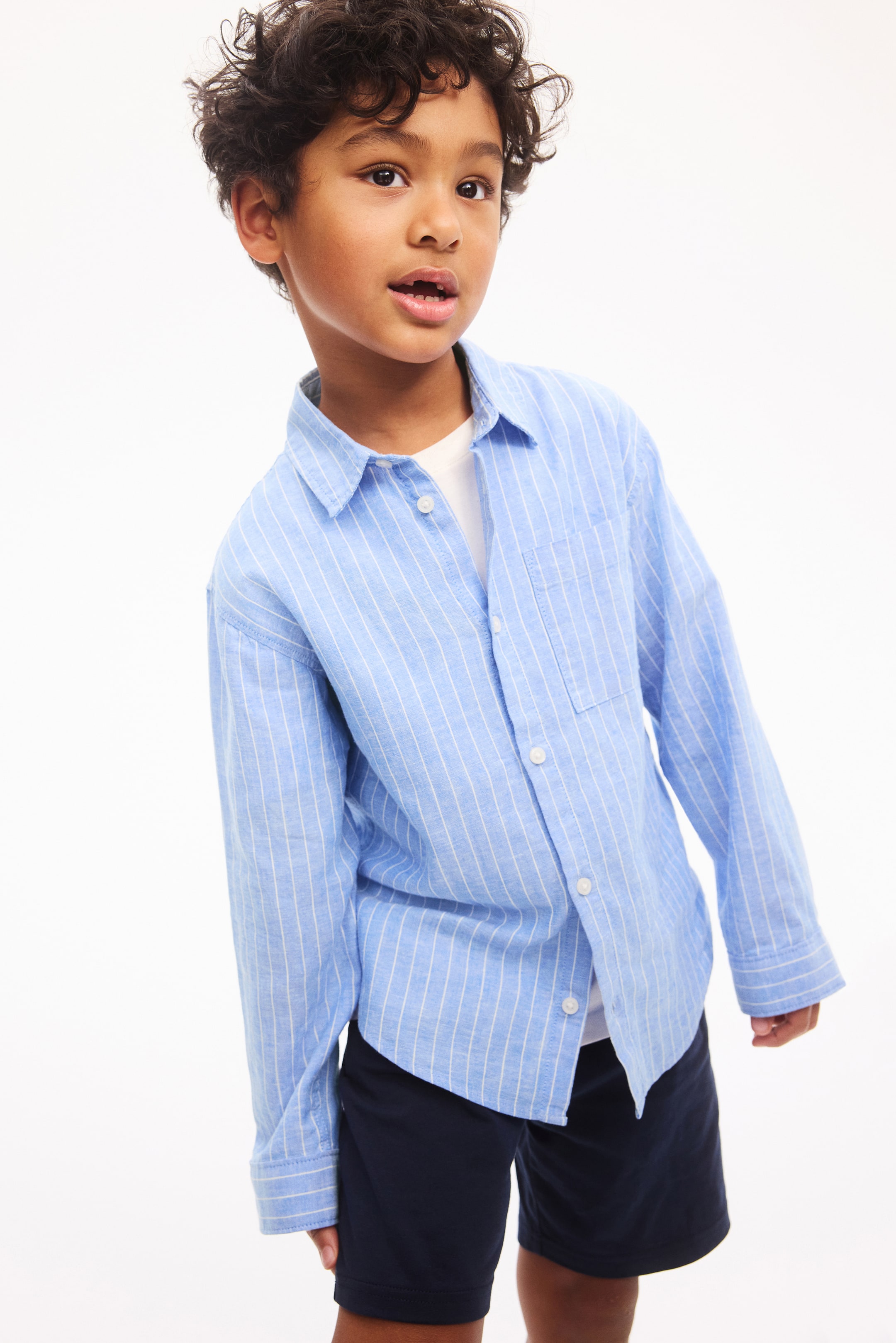 Ver imagem maior: Uma camisa azul‑claro de mangas compridas com finas riscas verticais brancas apresenta uma gola pontiaguda, abertura frontal com botões brancos e um único bolso de remendo no lado esquerdo do peito. Está usada aberta sobre uma t-shirt branca.