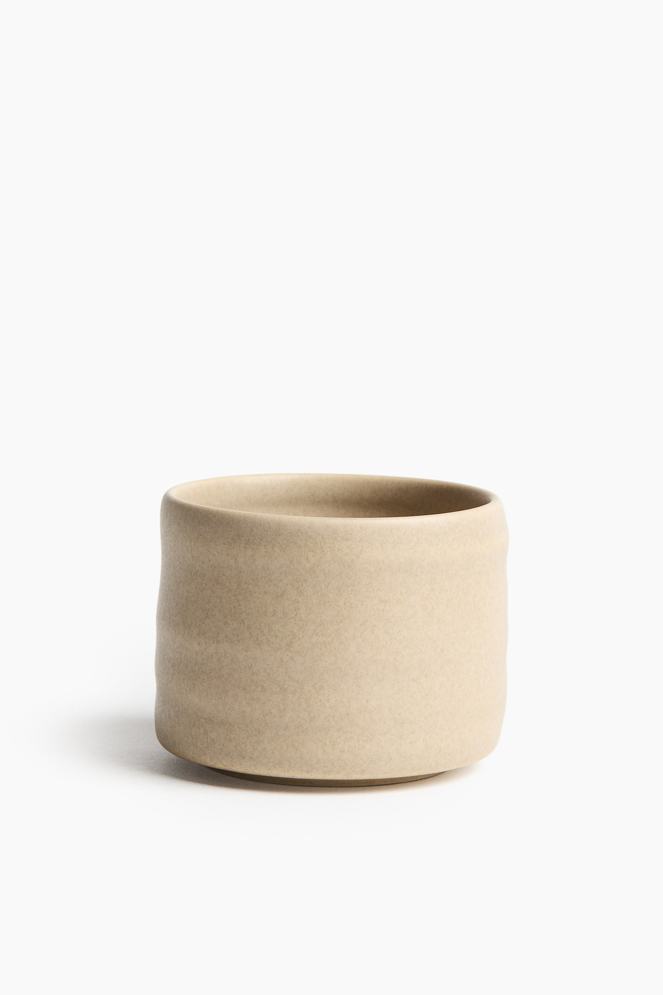 Grotere afbeelding bekijken: Een lichtbeige keramische pot staat en is licht naar rechts gericht. Zijn cilindrische vorm heeft subtiel onregelmatige zijkanten, en een gladde, afgeronde rand vormt de bovenkant van de opening.
