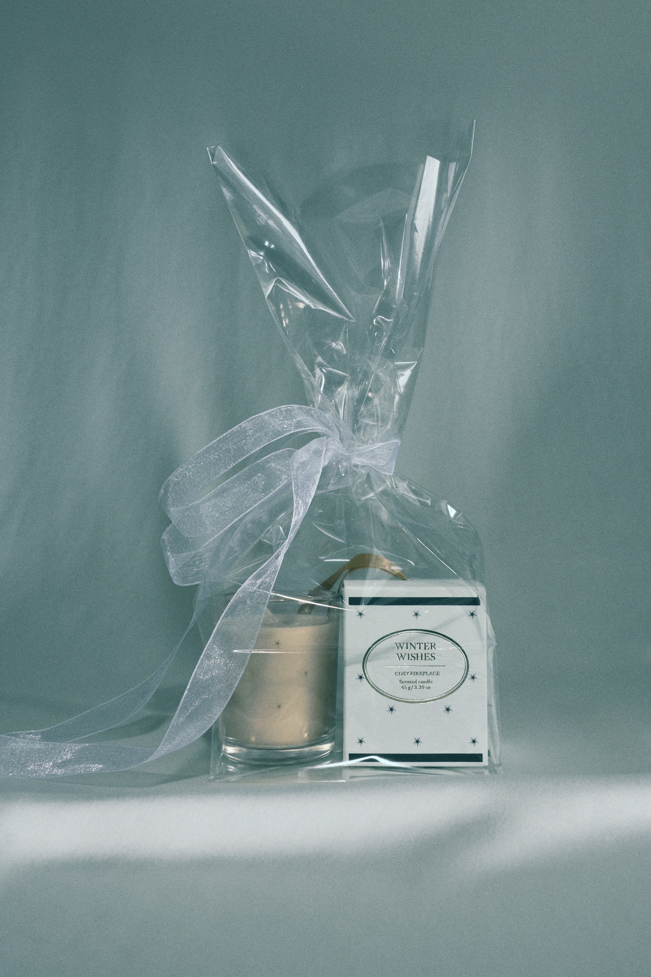 Ampliar la imagen: Una vela aromática en un portavelas de vidrio transparente decorado con estrellas doradas está junto a su caja de empaque blanca, que presenta un lazo dorado para colgar y el texto "Winter Wishes". Ambos artículos están empaquetados juntos dentro de una bolsa de regalo de celofán transparente, amarrada con un listón blanco translúcido.