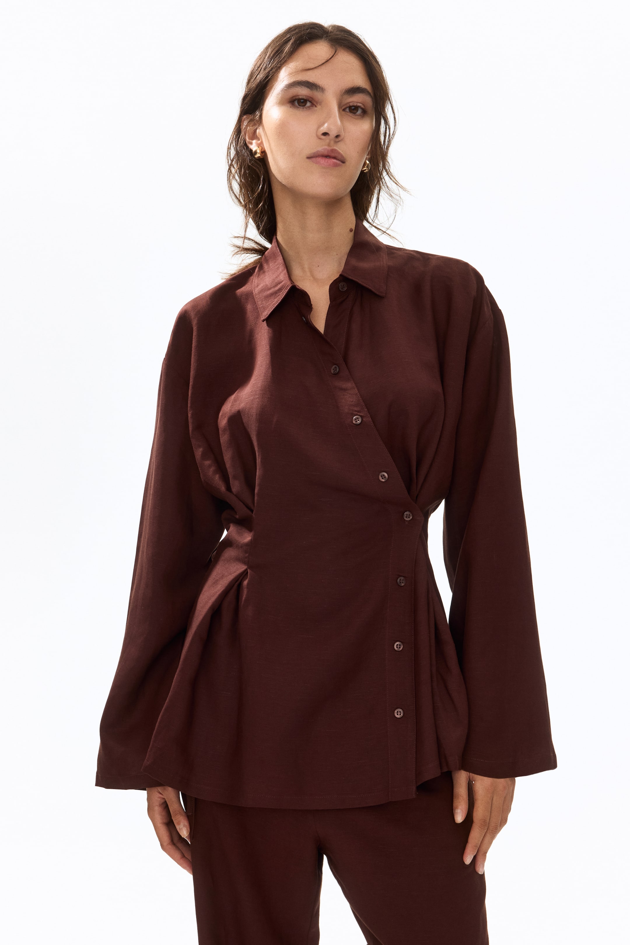 View larger image: The model is showcasing a deep brown shirt featuring a double-layered collar, dropped shoulders, and full-length sleeves. This piece is designed with a wrapover front, highlighted by an angled button band and box pleats at the waist, offering a cinched-in silhouette. It is paired with coordinating deep brown trousers.