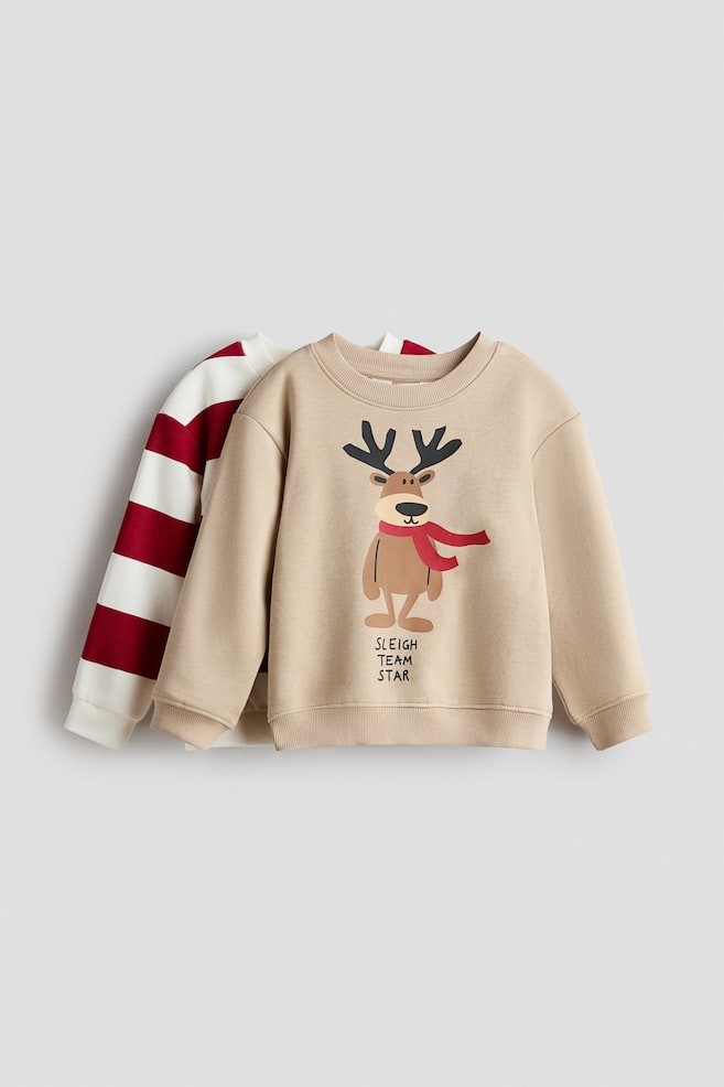 A beige long-sleeve sweater is shown from the front, layered over a red and white striped sweater. The beige sweater features a round rib-trimmed neckline, dropped shoulders, ribbed cuffs and hem, and a printed cartoon reindeer graphic with the text "SLEIGH TEAM STAR" on the chest. - 1