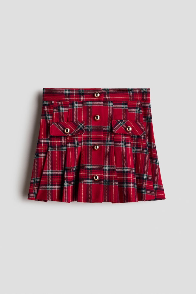 A front-facing red tartan pleated mini skirt featuring a vertical row of four decorative gold-tone dome buttons down the front and two symmetrical flap pockets, each accented with a matching gold button. The skirt showcases a structured waistband and flared knife pleats along the lower half. - 1