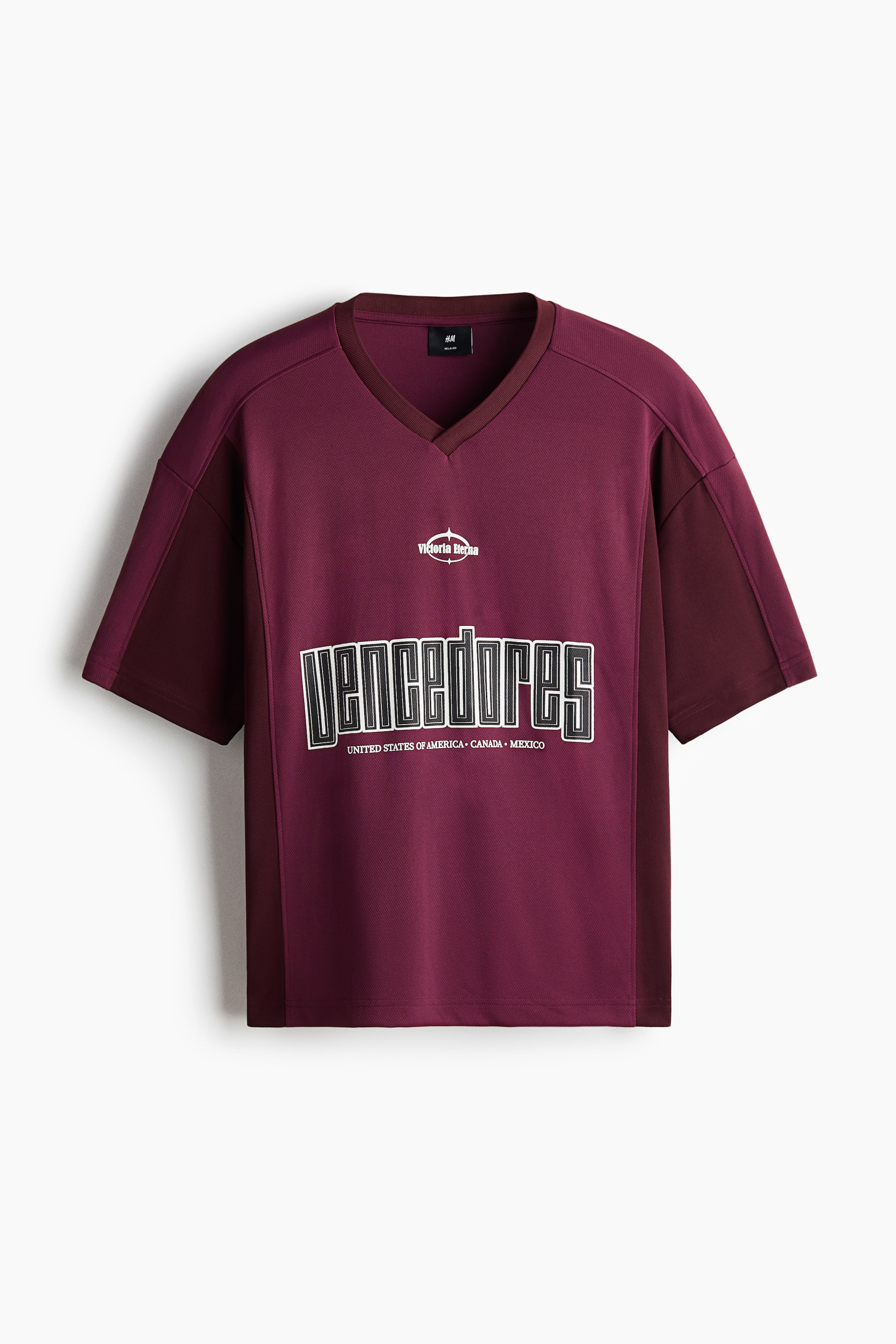 View larger image: A maroon T-shirt is shown from the front, featuring a subtle diagonal knit texture, a darker ribbed V-neckline, and short sleeves with darker side panels. It is displaying 'Victoria Berna' and 'VENCEDORES' with 'UNITED STATES OF AMERICA • CANADA • MEXICO' print motifs on its chest, presenting a relaxed fit and a straight-cut hem.