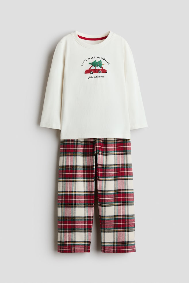 A front-facing pyjama set featuring a long-sleeved, off-white jersey top with a round neckline and a graphic of a red car carrying a green tree on the chest. The top is paired with straight-leg flannel pants in a red, green, and cream plaid pattern. - 1