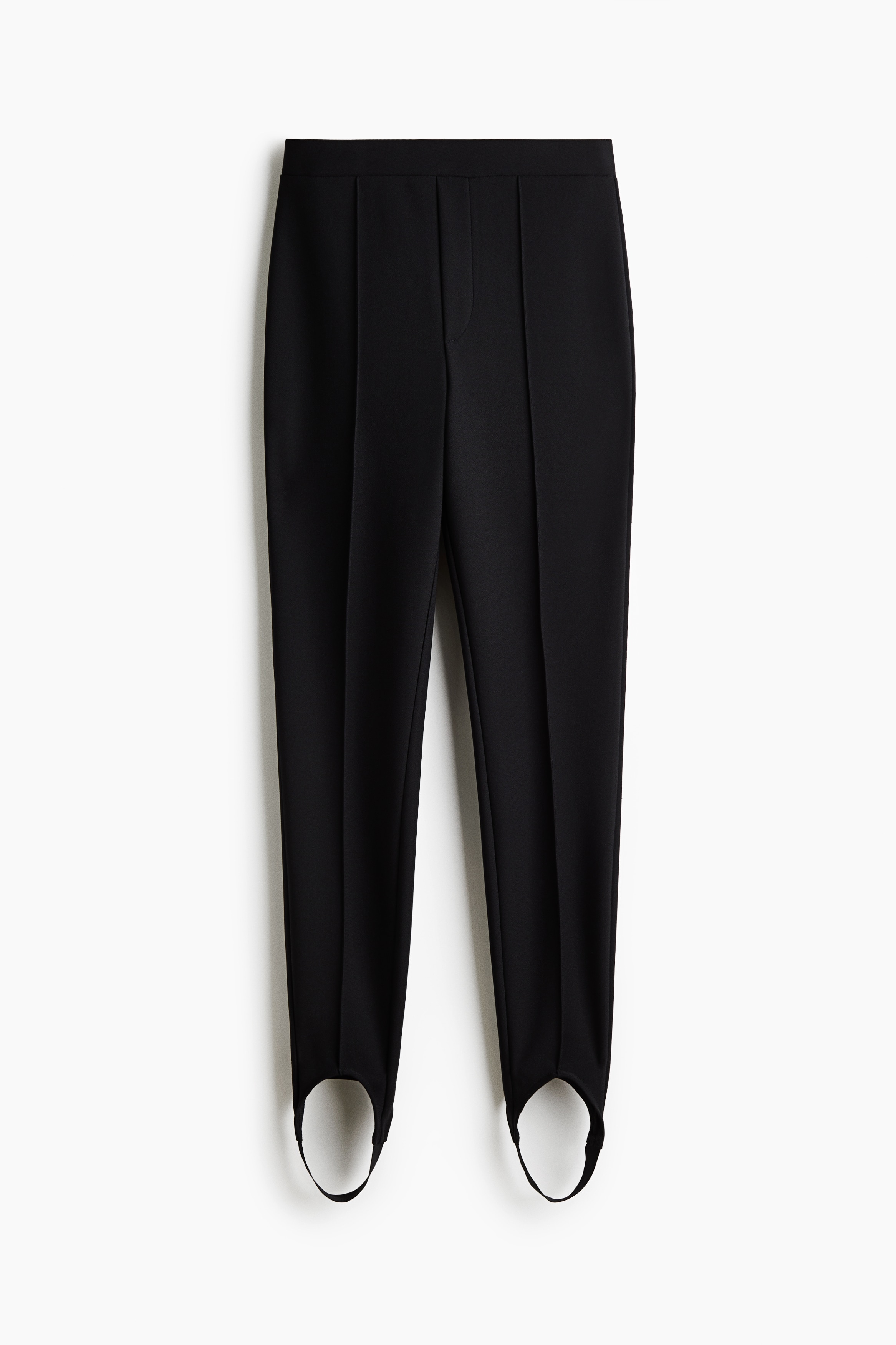 The Crease Front Stirrup Leggings in dark navy are being worn by the model, featuring a sharp vertical seam down the centre of each leg and stirrup straps looped under the feet. The leggings are styled with a layered black high-neck top, a navy blue button-down shirt, an oversized olive trench coat, and black high-heeled shoes.