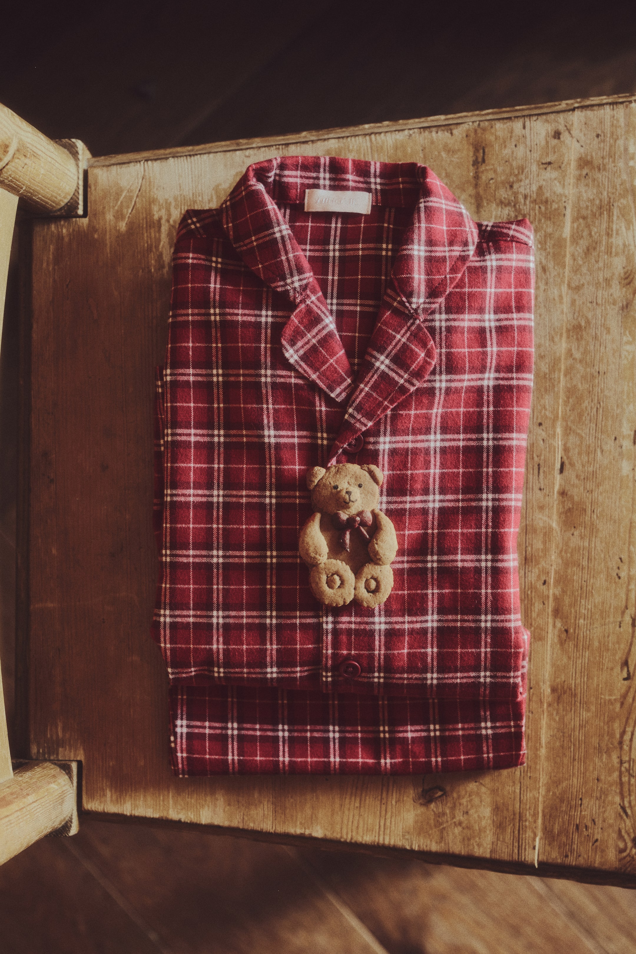 Grotere afbeelding bekijken: Een set rode pyjama's met ruitjespatroon ligt opgevouwen en van voren zichtbaar, met een overhemd met knopen, revers met inkeping en bijpassende broek. Een klein koekje in de vorm van een teddybeer rust op het midden van de voorkant van het overhemd.