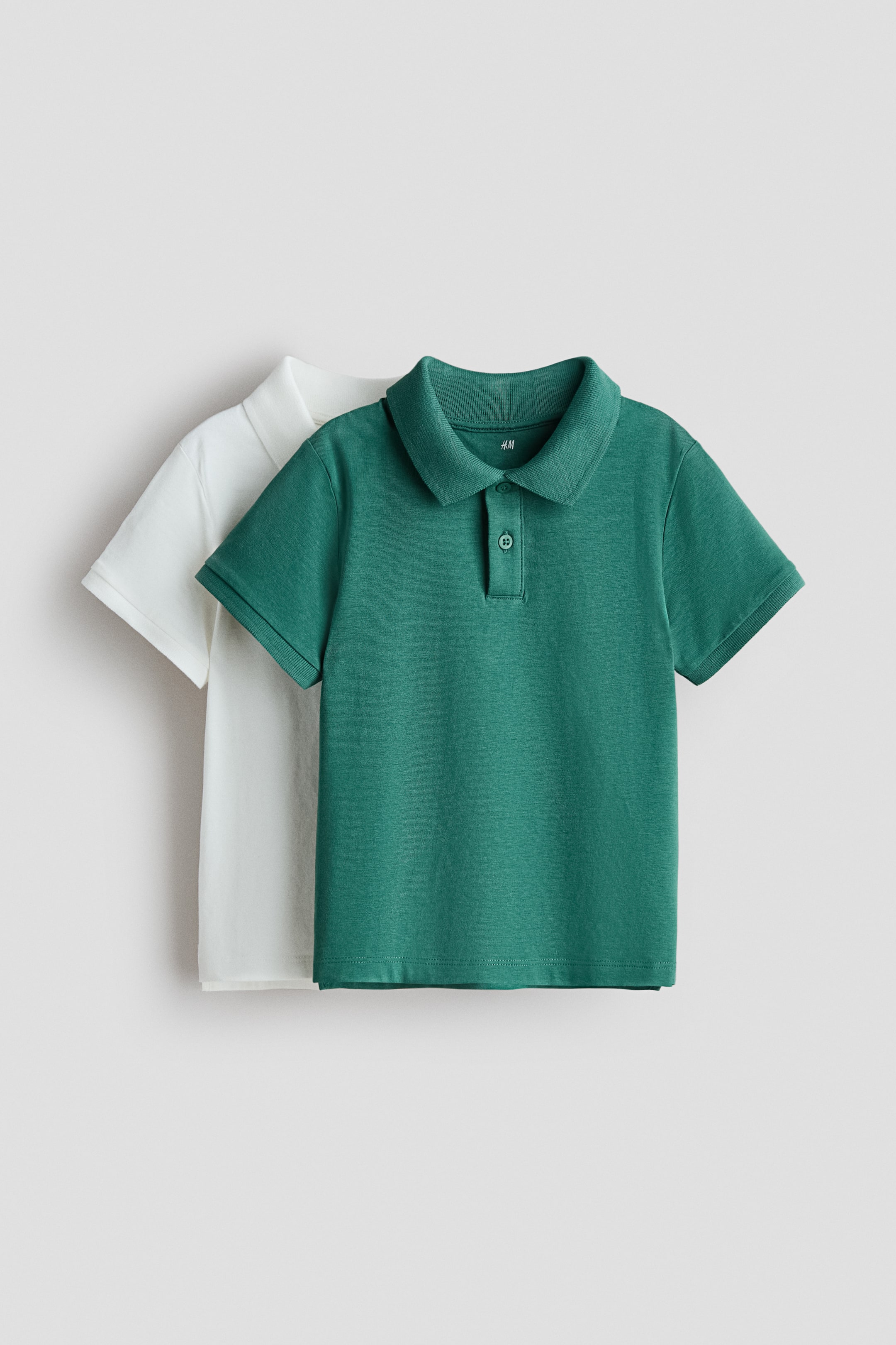 View larger image: Two polo shirts are showcased from the front, with a dark green shirt layered over a white one. The front dark green polo shirt features a ribbed collar and a two-button placket with matching buttons, while both shirts display short sleeves with ribbed cuffs.