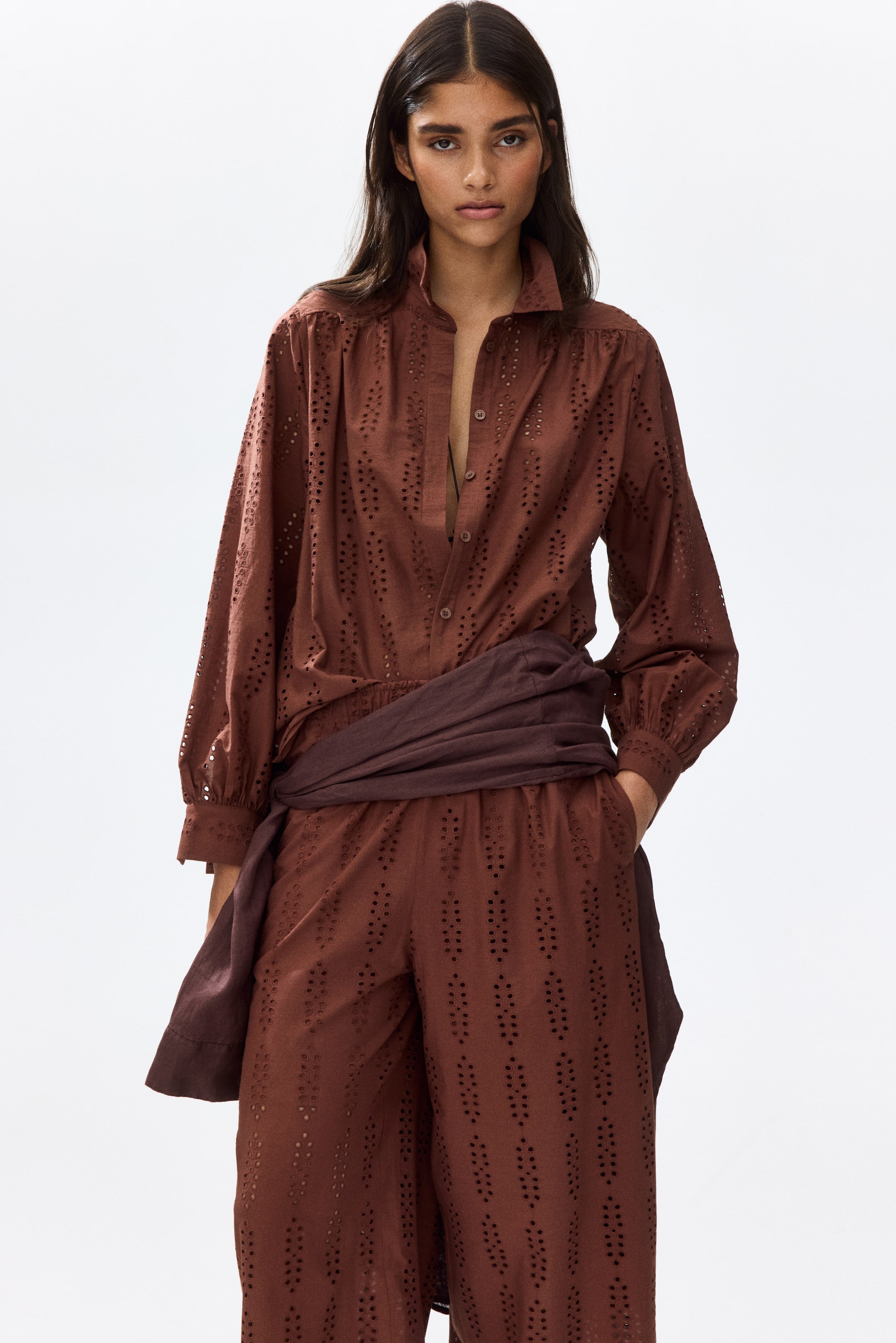 View larger image: The model is showcasing a relaxed-fit broderie anglaise shirt in a rich brown hue. It features a collar with a visible button, a partially unbuttoned concealed front placket, and a gathered yoke that gracefully extends over the shoulders. The shirt is completed with long balloon sleeves that have buttoned cuffs. Complementing the shirt, the model is also wearing matching broderie anglaise pants. To accentuate the ensemble, a dark brown sash is elegantly wrapped around the waist.