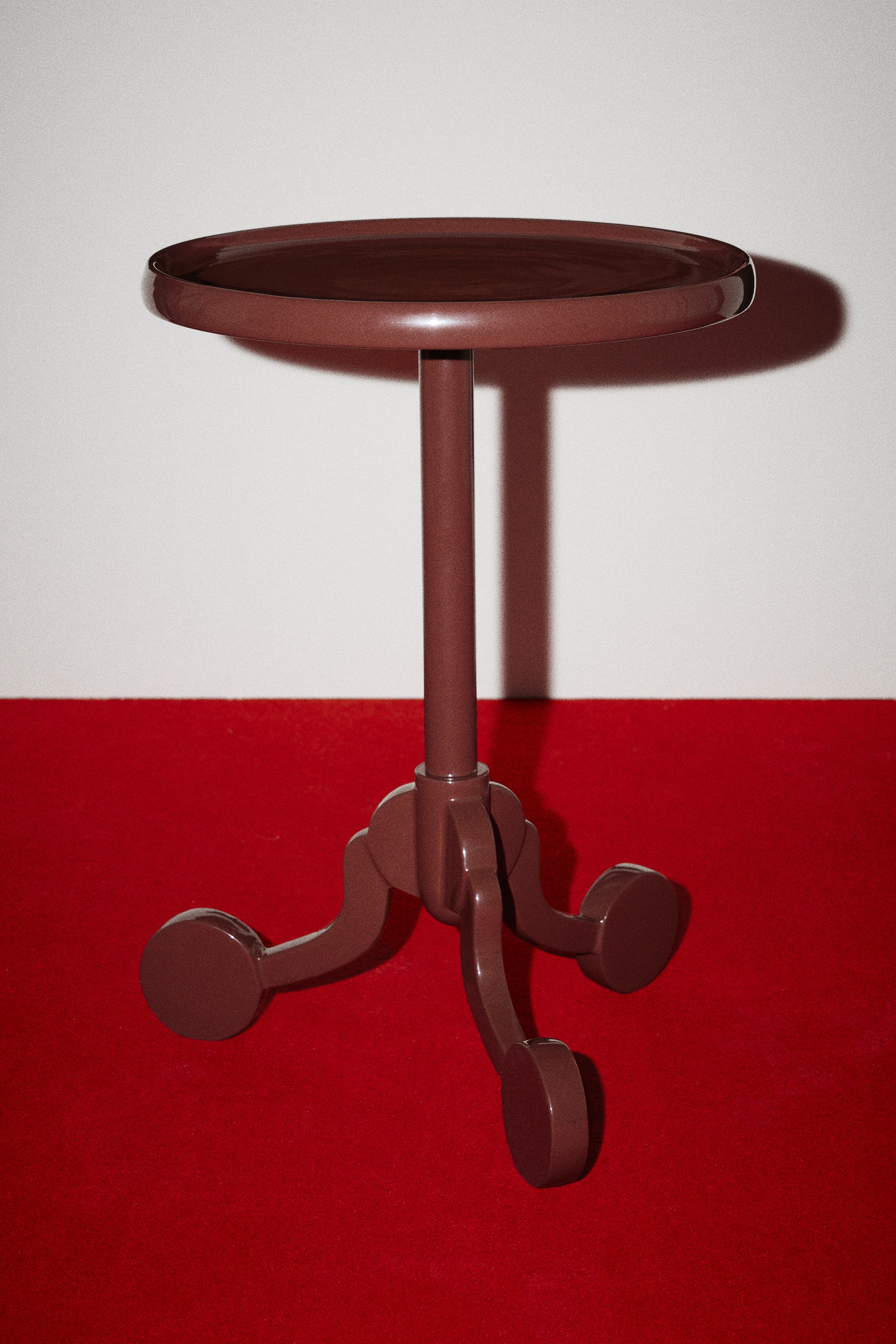 A glossy, dark reddish-brown side table featuring a round, tray-like top with a raised edge. The central column supports the top, connecting to a three-legged base with curved legs, each terminating in a round foot.
