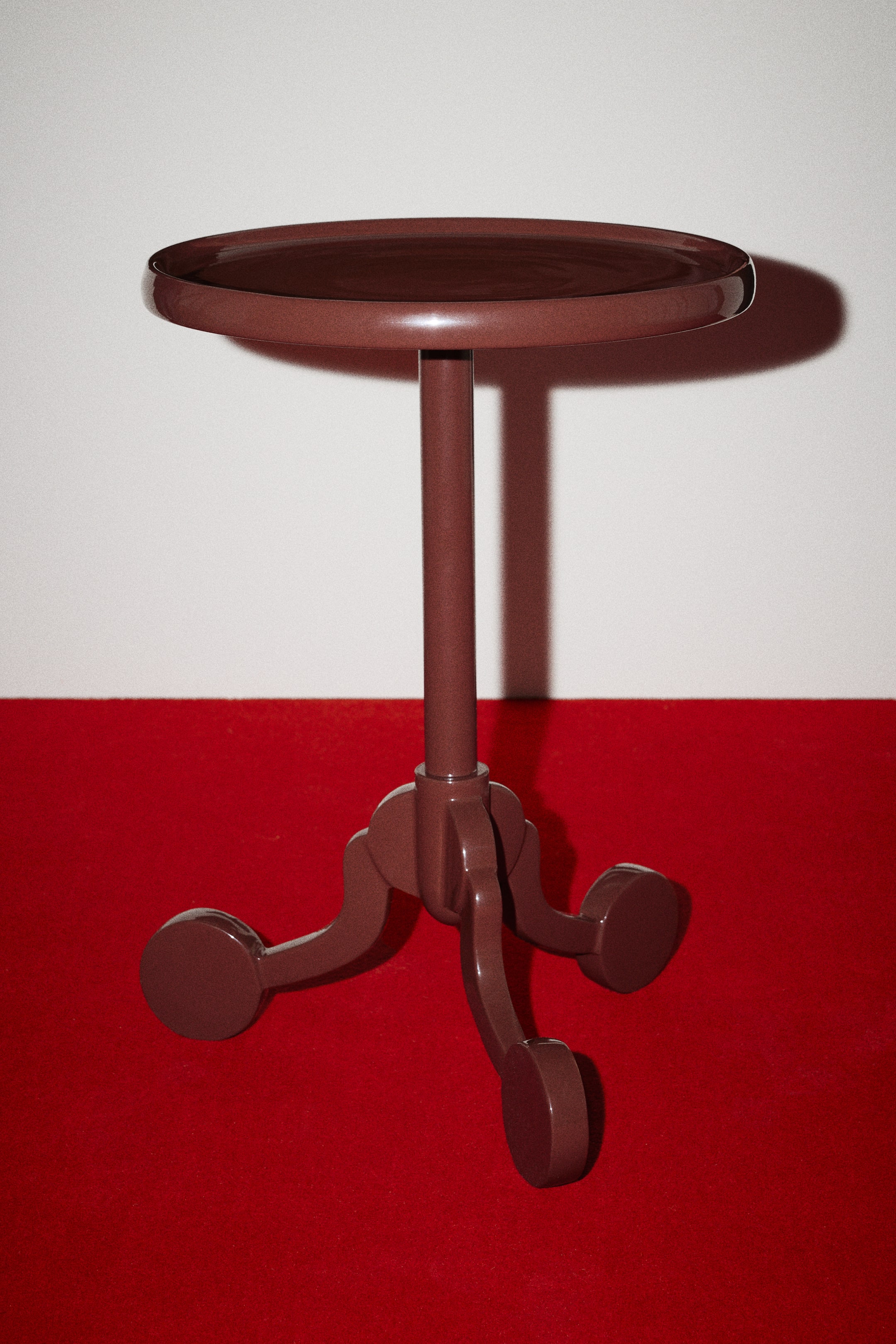 View larger image: The Myra side table is shown from a front-facing angle, highlighting its glossy, dark brown lacquered finish. It features a circular, tray-like tabletop with a raised rim, supported by a central cylindrical stem extending down to a tripod base with three curved legs ending in flat, circular feet.