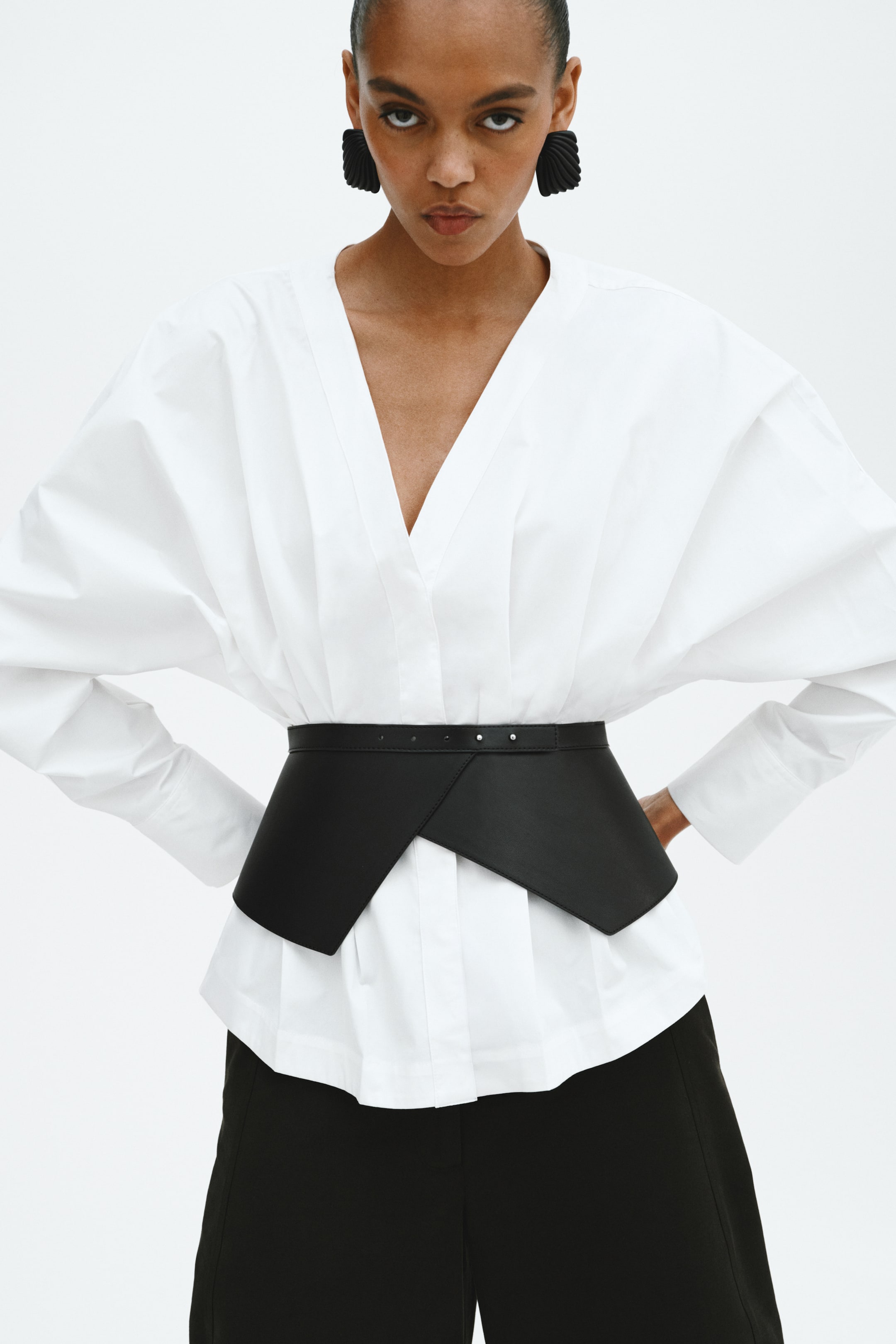 View larger image: A black structured wide belt cinches the waist over a white shirt, showcasing an asymmetrical angular front design. Small silver-toned studs detail the top edge of the belt.