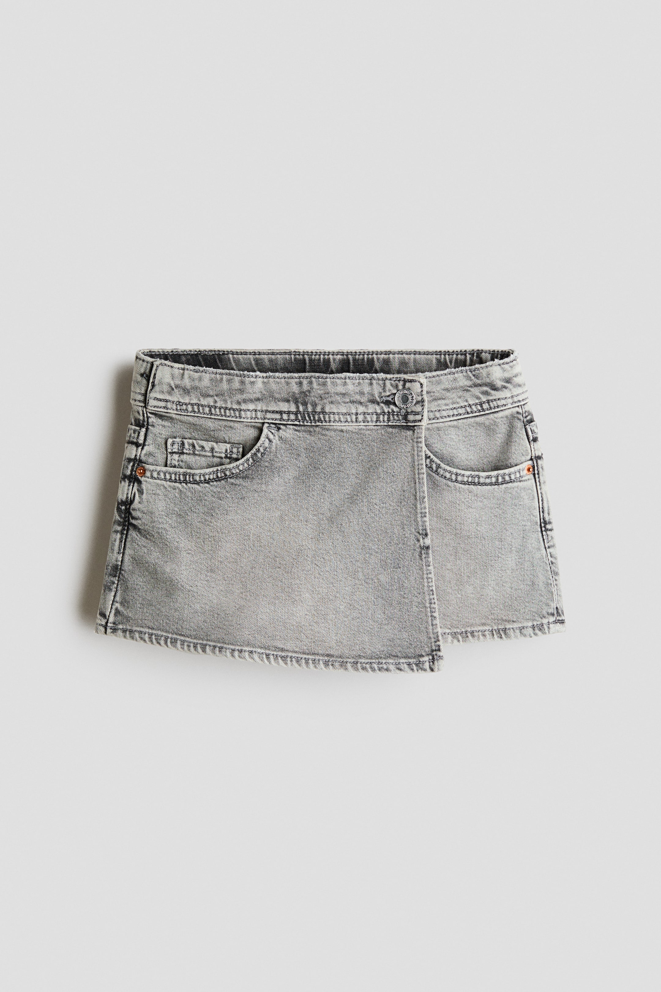 View larger image: A washed grey denim skort is showcased from the front, boasting an elasticated waistband and two curved front pockets with a coin pocket. It features a wrapover front with a single button closure and a distressed hem.