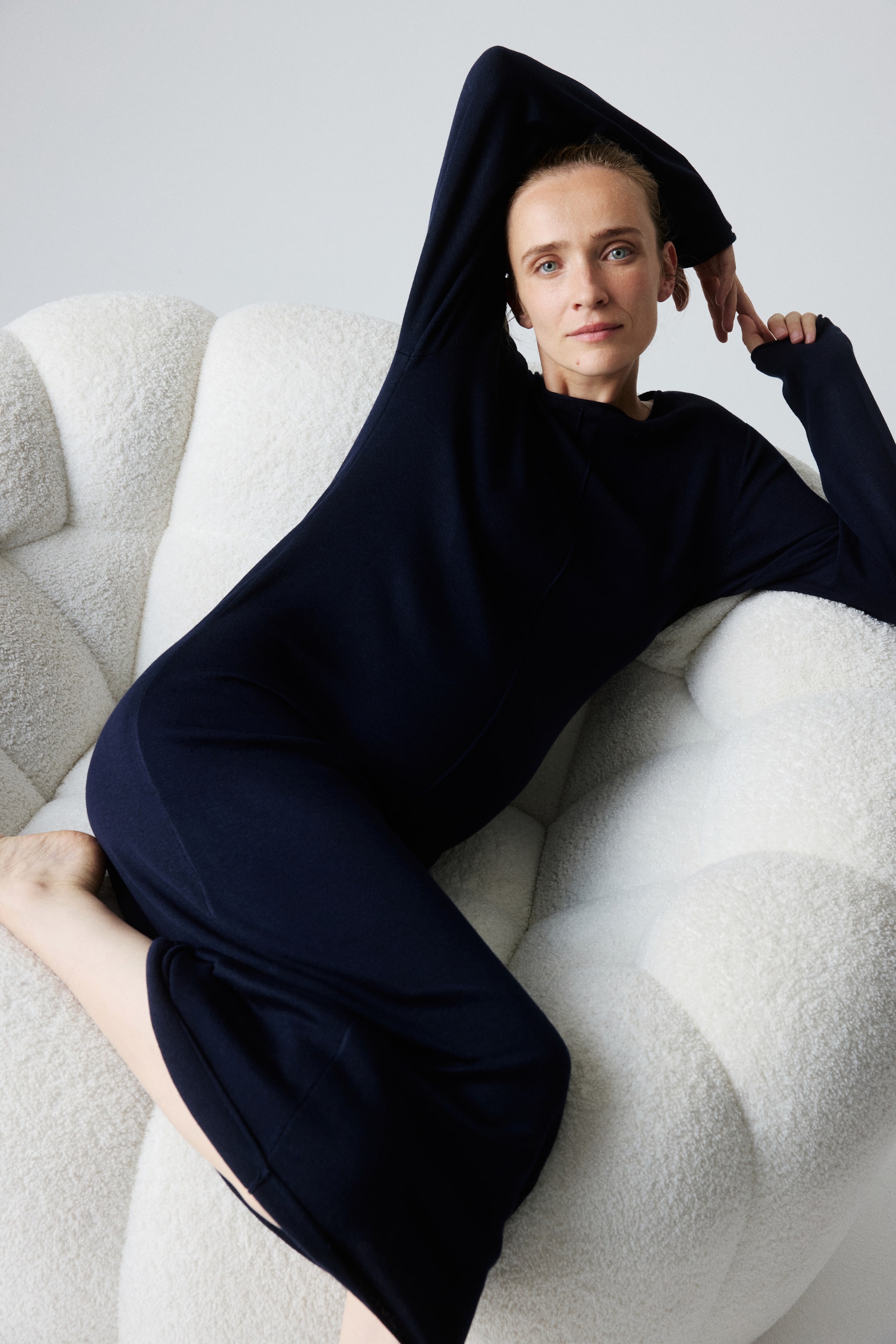 Преглед на по-голямо изображение: Моделът е облечен в тъмна, прилепнала трикотажна рокля до прасеца с лодковидно деколте и дълги ръкави. Роклята е с открит шев по предната част и се спуска свободно около тялото ѝ, докато тя е в полулегнало положение.