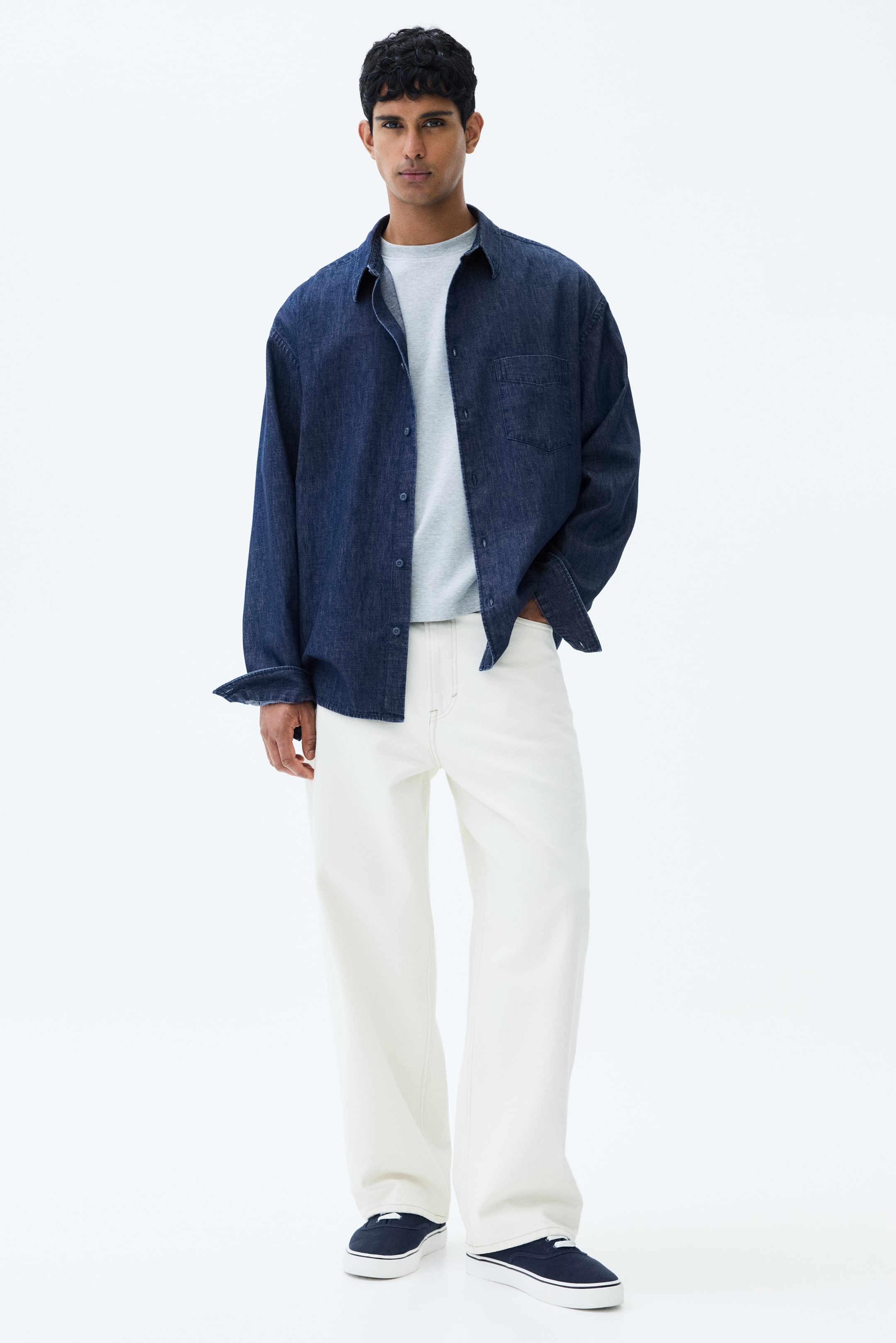 View larger image: The model is showcasing a dark blue rigid cotton denim shirt, styled open, featuring a collar, French front, and a patch chest pocket. It has long sleeves with visible cuffs and a subtly rounded hem, offering a relaxed fit over a light grey t-shirt.