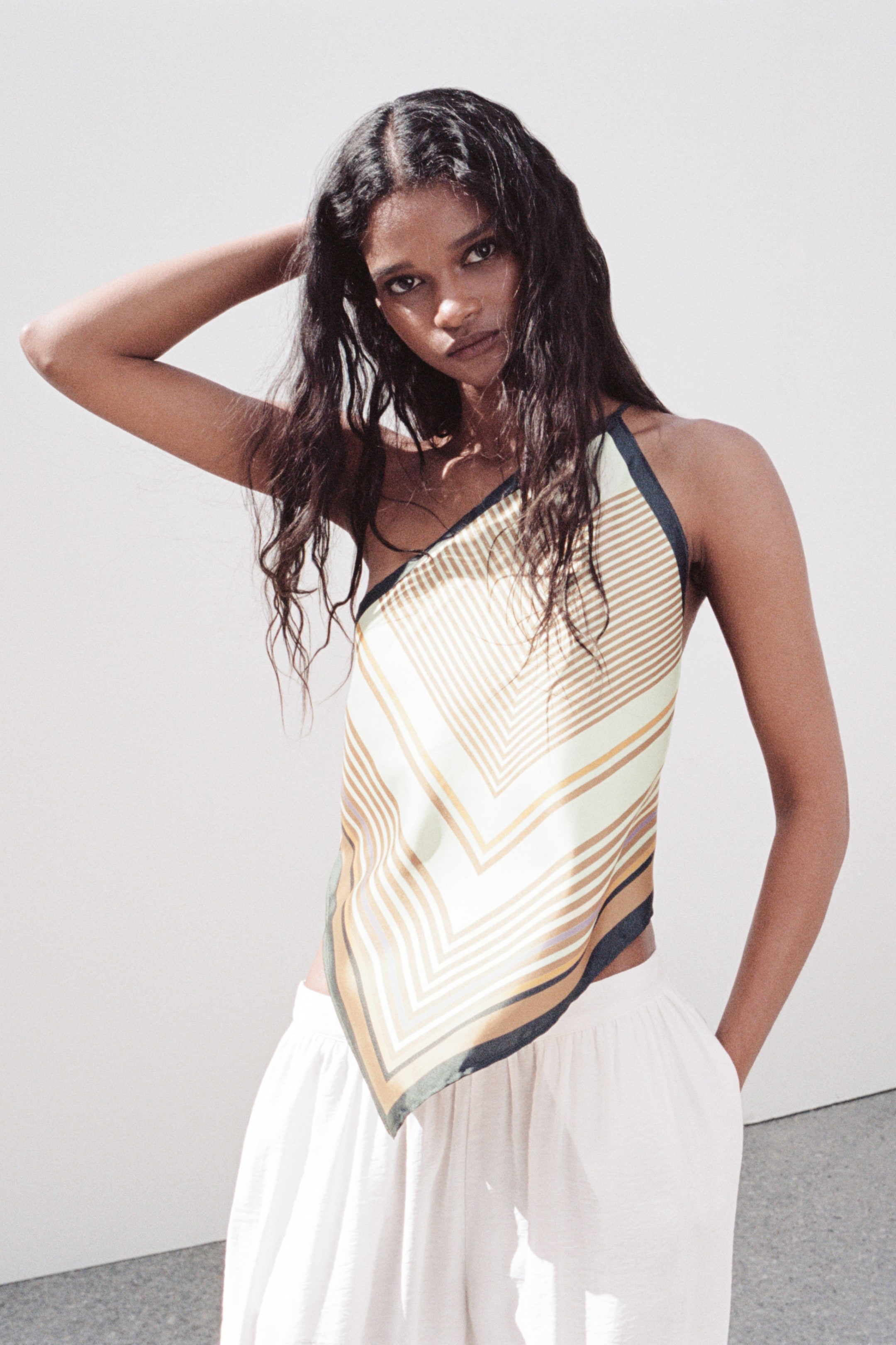 View larger image: A triangle bandana-style top features concentric V-shaped stripes in cream, gold, brown, and navy. Designed with a halter neck and a pointed hem that reveals the midriff, it's paired with loose white pants.