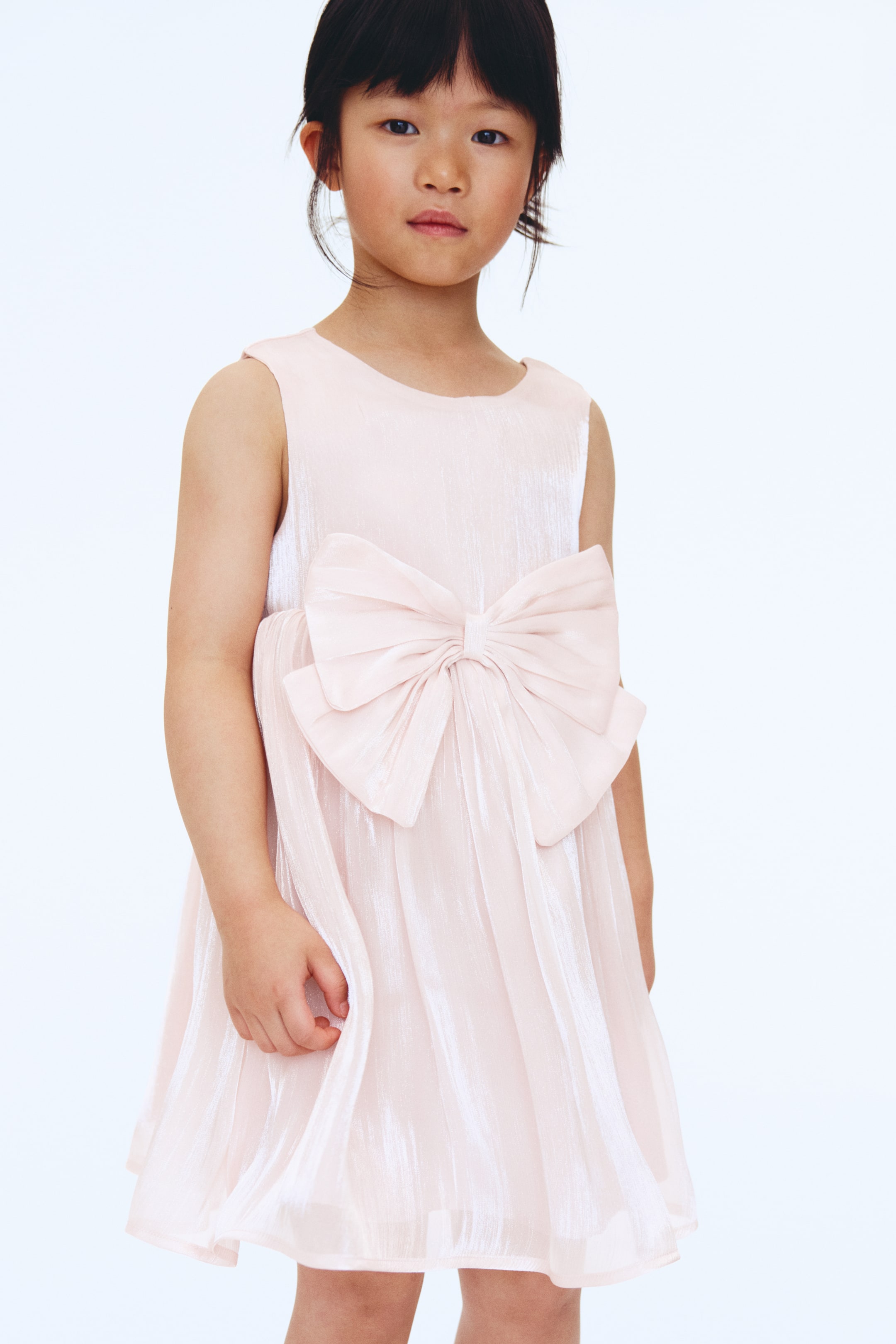 View larger image: A light pink sleeveless dress showcases a round neckline with a prominent bow accent at the front. This dress features a shimmering, crinkled fabric that flows into an A-line skirt.