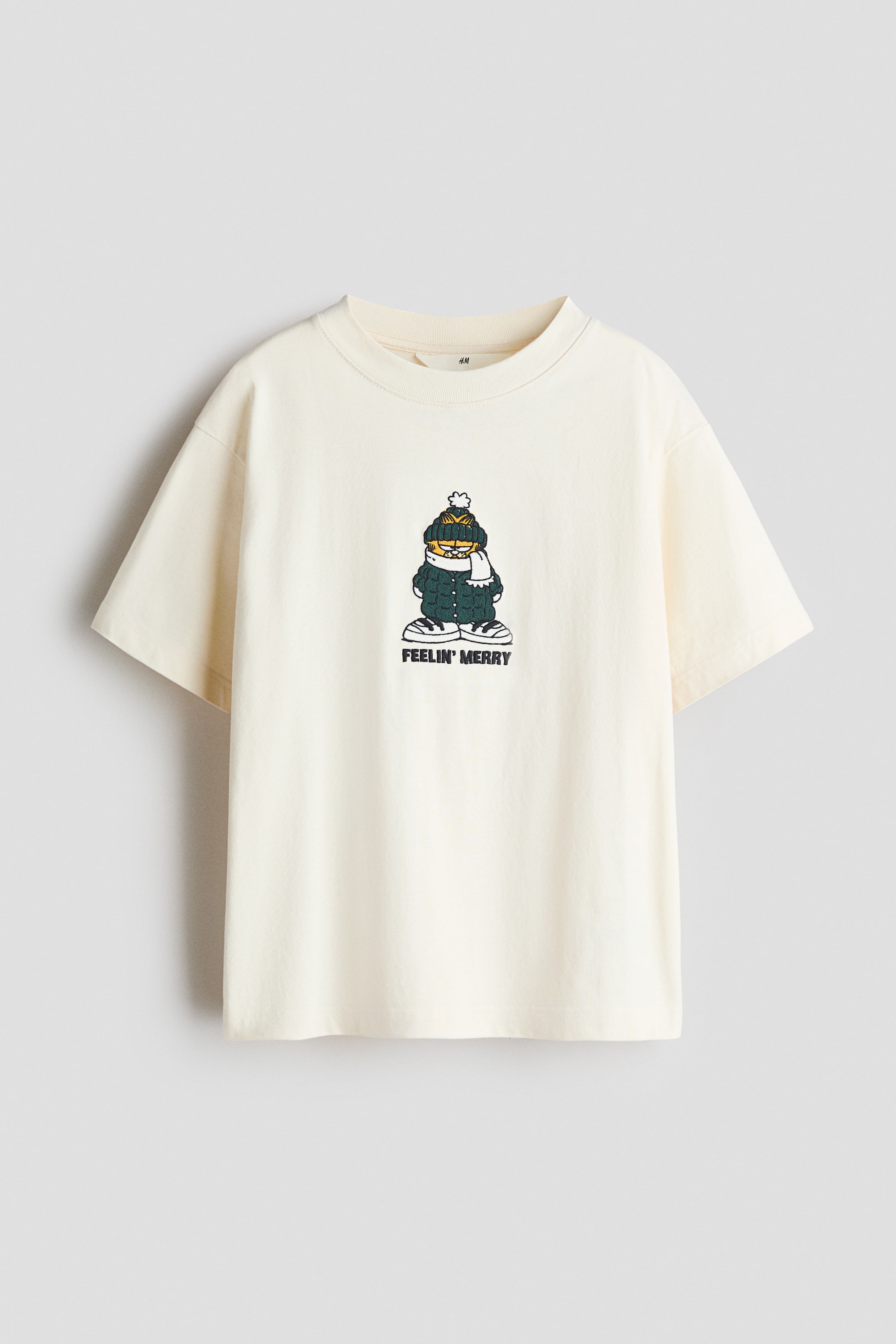 View larger image: An off-white cotton jersey T-shirt is shown from the front, featuring a round rib-trimmed neckline and dropped shoulders. The centre chest displays a printed graphic of a cartoon cat wearing winter clothing, with the text "FEELIN' MERRY" printed directly below it.