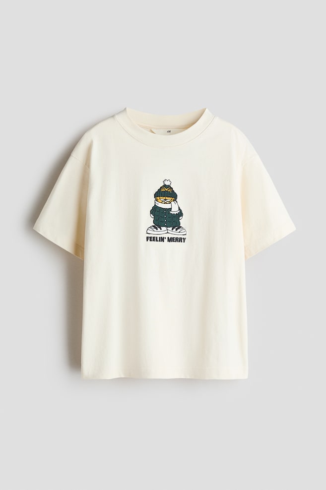 An off-white cotton jersey T-shirt is shown from the front, featuring a round rib-trimmed neckline and dropped shoulders. The centre chest displays a printed graphic of a cartoon cat wearing winter clothing, with the text "FEELIN' MERRY" printed directly below it. - 1