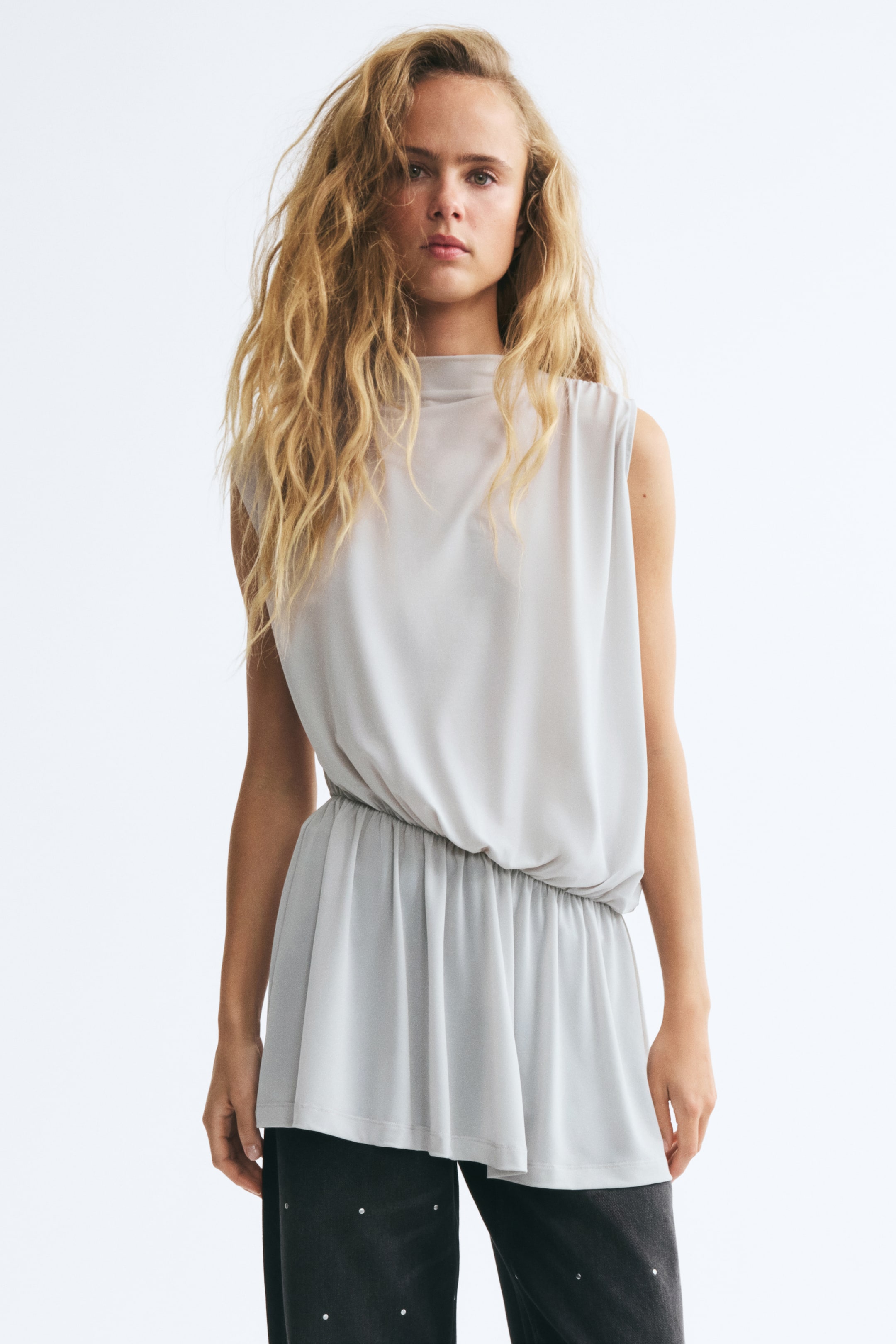 View larger image: The model is wearing a light gray, sleeveless, loose-fit draped top with a high neckline and an asymmetrical gathered waist for a draped effect. The top falls into a flared hemline and is styled with dark, embellished trousers.